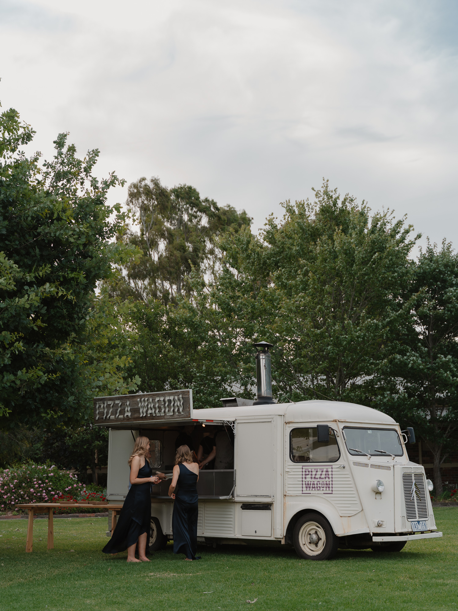 Two women in formal dresses order at a white pizza food truck named 'Pizza Wagon' on a grassy lawn with trees in the background. Pizza Wagon Wedding catering