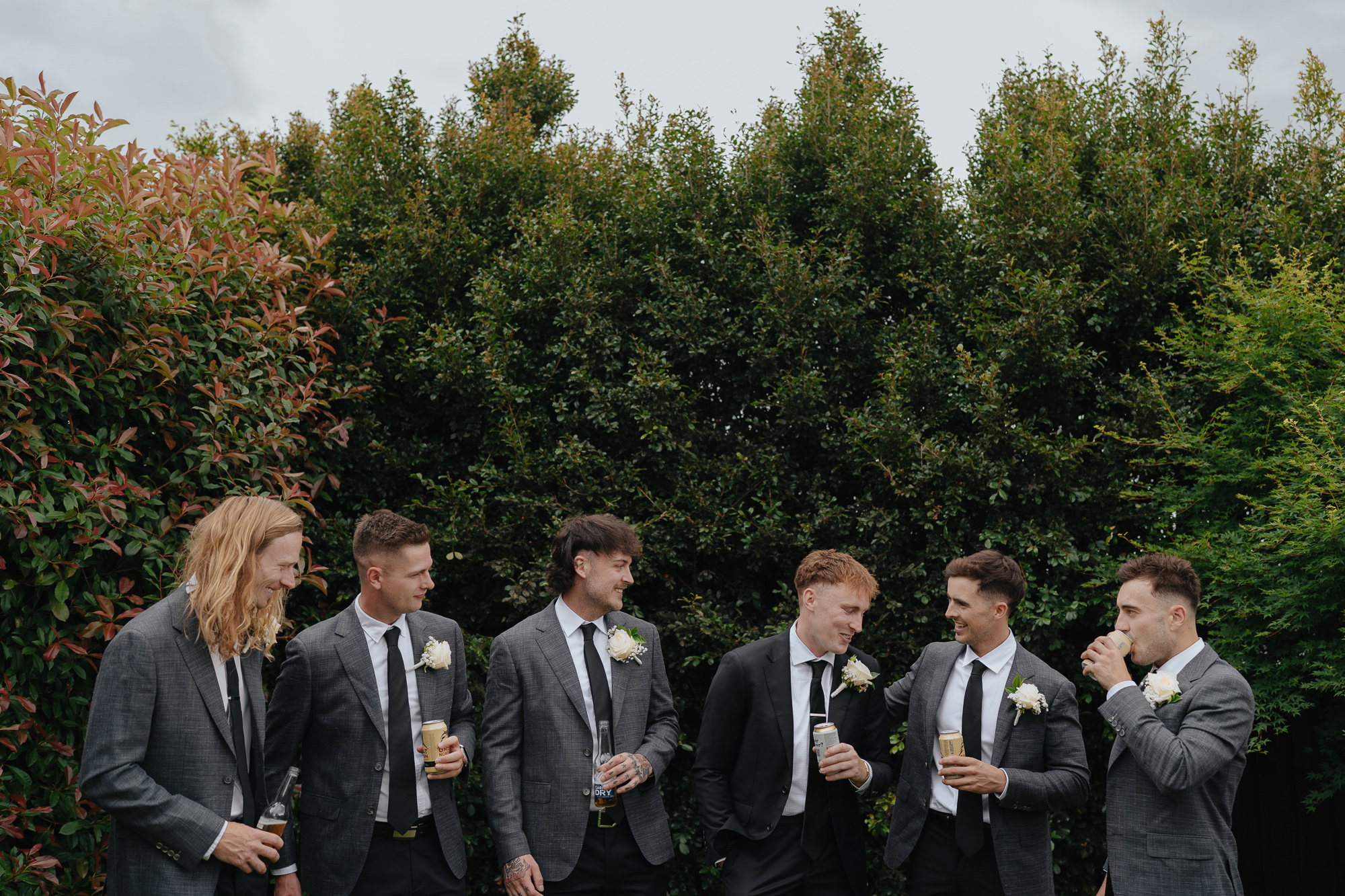 Group of seven men in suits, some with boutonnières, at an outdoor event, holding drinks and engaging in conversation, with green foliage and trees in the background.