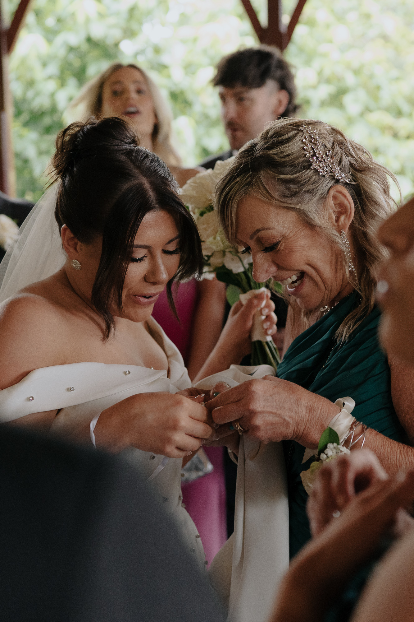 A bride and an older woman, likely her mother, sharing a joyful moment during a wedding at an outdoor setting. The bride is wearing a white wedding gown with off-shoulder sleeves, and the woman is holding the bride's hand, both smiling.