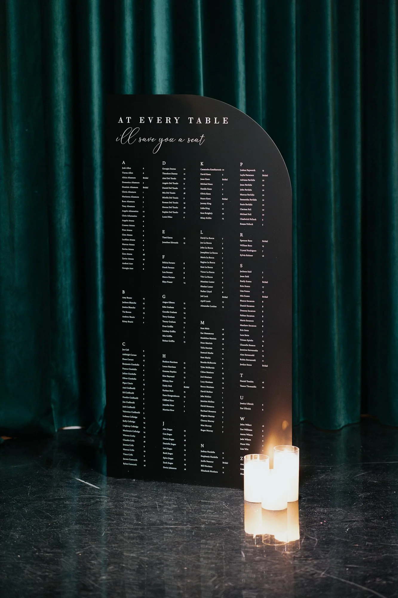 A black seating chart with white text titled 'At Every Table, I'll Save You a Seat', placed on a stand in front of green curtains, with two lit candles at the base.
