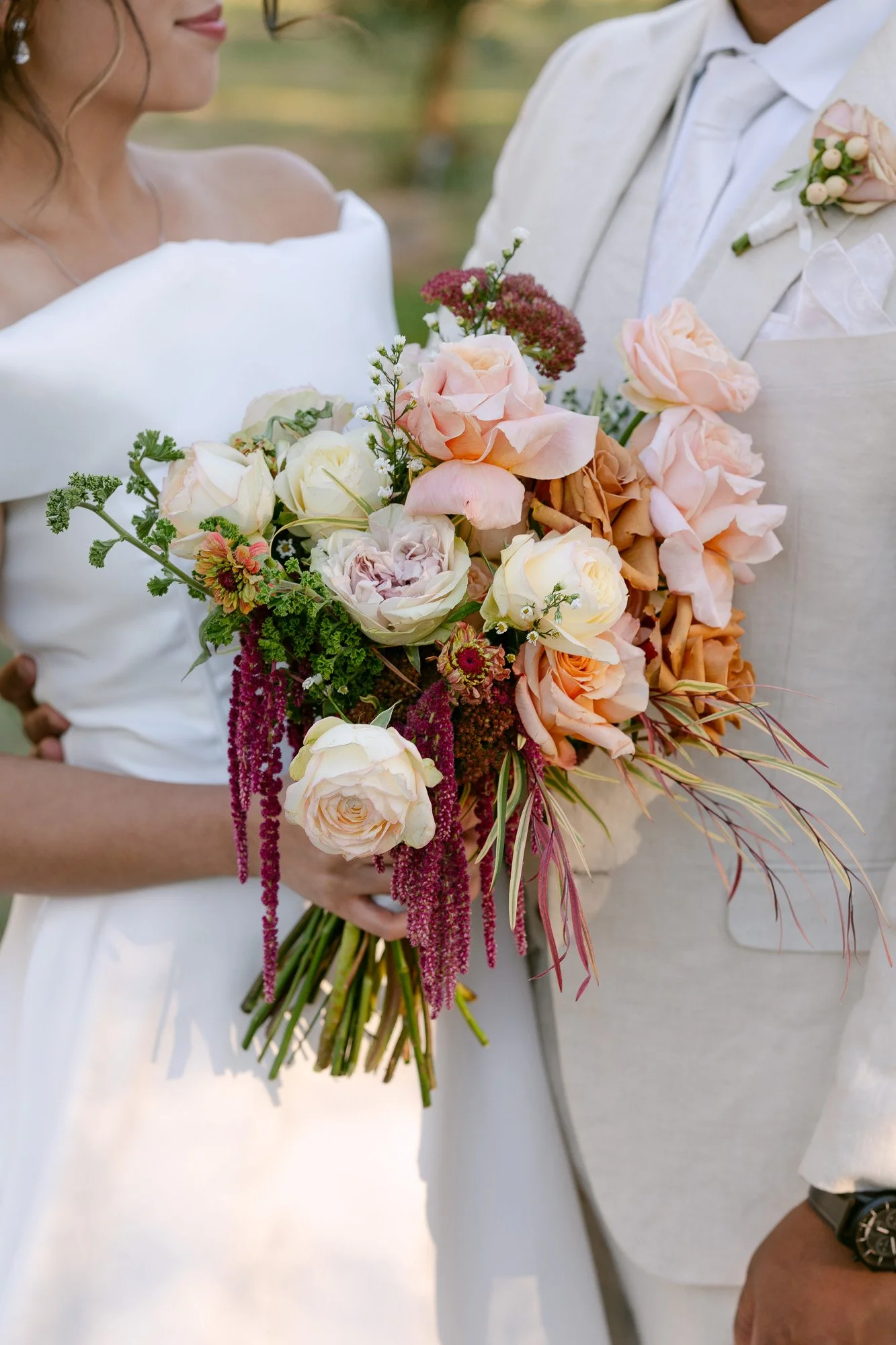 A bride and groom holding a bouquet of pink, white, and peach roses, along with various greenery and accents, during their wedding.