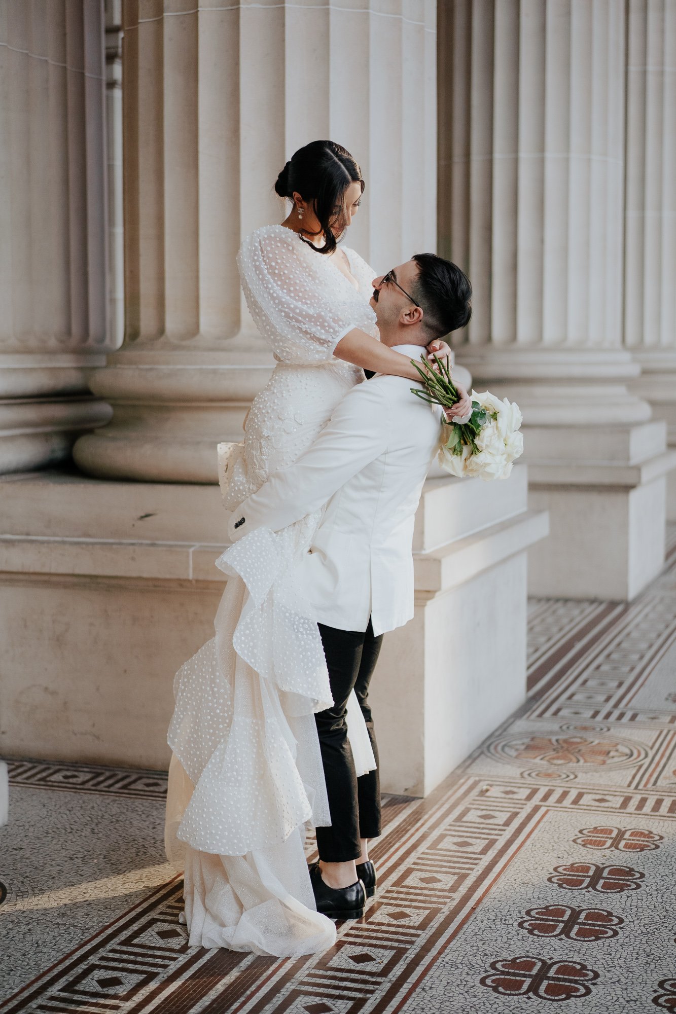 A couple dressed in wedding attire, the woman in a white wedding gown and the man in a white suit jacket, sharing a romantic moment inside a grand building with tall columns and intricate tile flooring.