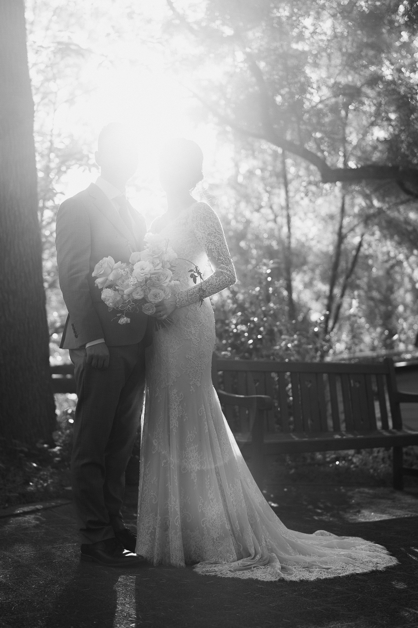 A black and white photo of a bride and groom standing outdoors with sunlight shining behind them. The bride is holding a bouquet of flowers and wearing a lace wedding dress, while the groom is dressed in a suit. Chateau Wyuna