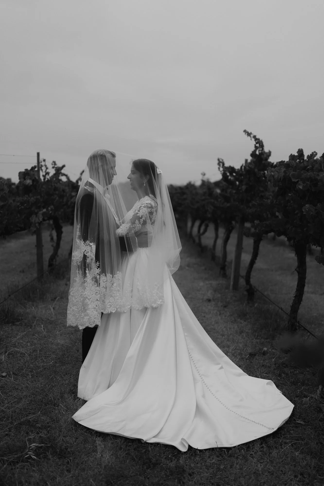 A black and white photo of a bride and groom standing close together in a vineyard at Witchmount Estate Functions & Restaurant, both wearing wedding dresses with the bride's train visible, and a veil covering both their faces.