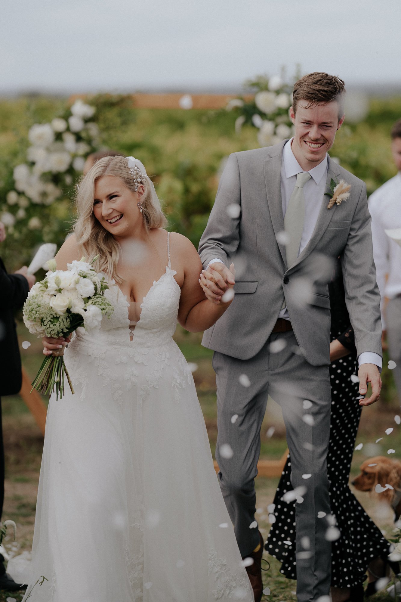 A newlywed couple holding hands and smiling during their outdoor wedding celebration at The Vines on Avon, surrounded by falling confetti and floral decorations.