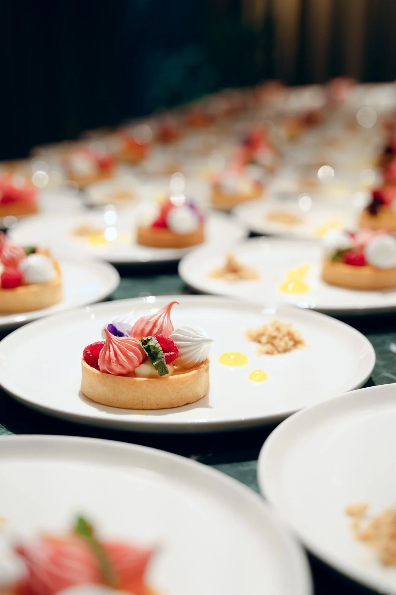 Multiple plates of desserts, with the front plate featuring a tart topped with berries, whipped cream, and decorative meringue, on a dark table.