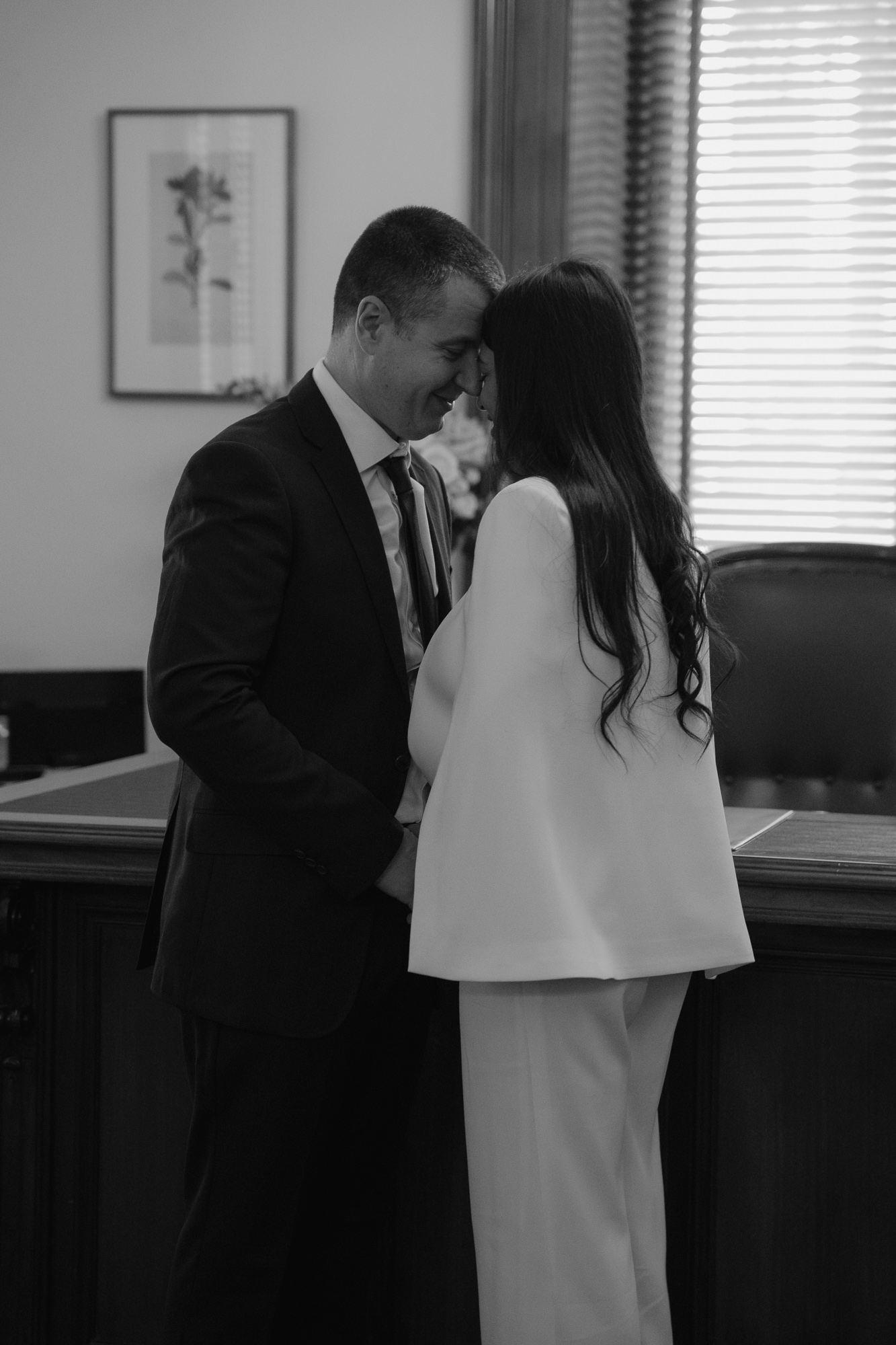 A man and woman with foreheads touching, smiling in a formal office setting, during a special moment. Melbourne Treasury Elopement Photography.