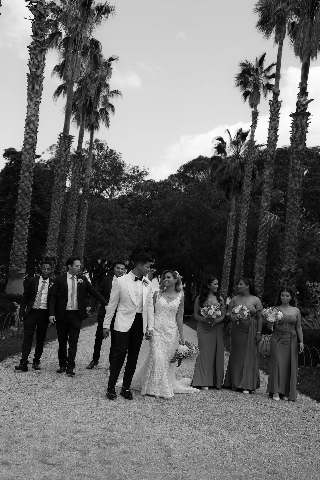 A black and white photo of a wedding party walking outdoors on a path lined with palm trees. The bride and groom are in the center, smiling and holding hands, surrounded by bridesmaids with bouquets and groomsmen at Williamstown Botanic Gardens.
