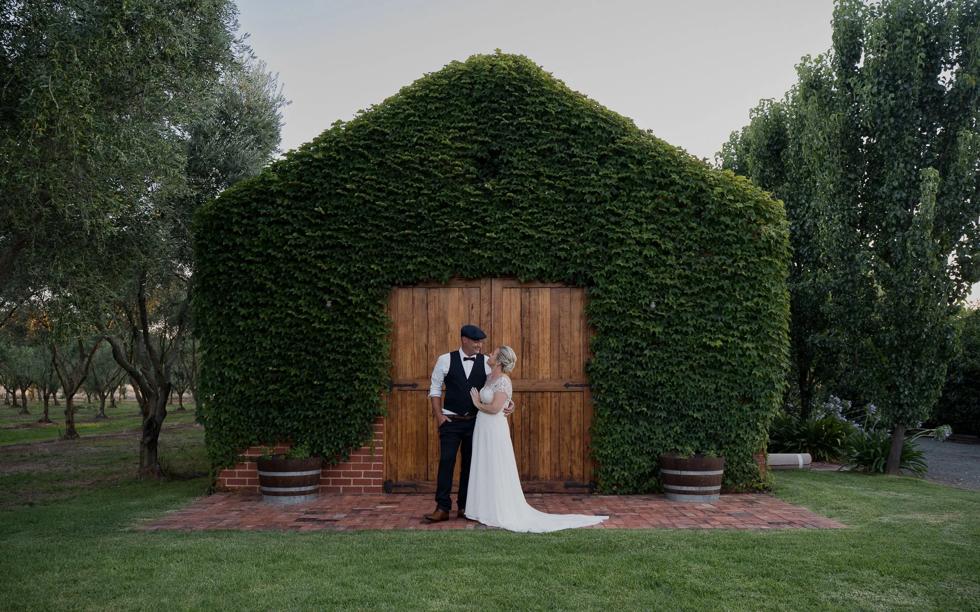 Bride and groom standing together in front of a large closed wooden door covered with green vines, outdoors on a brick patio, with trees and grass in the background.