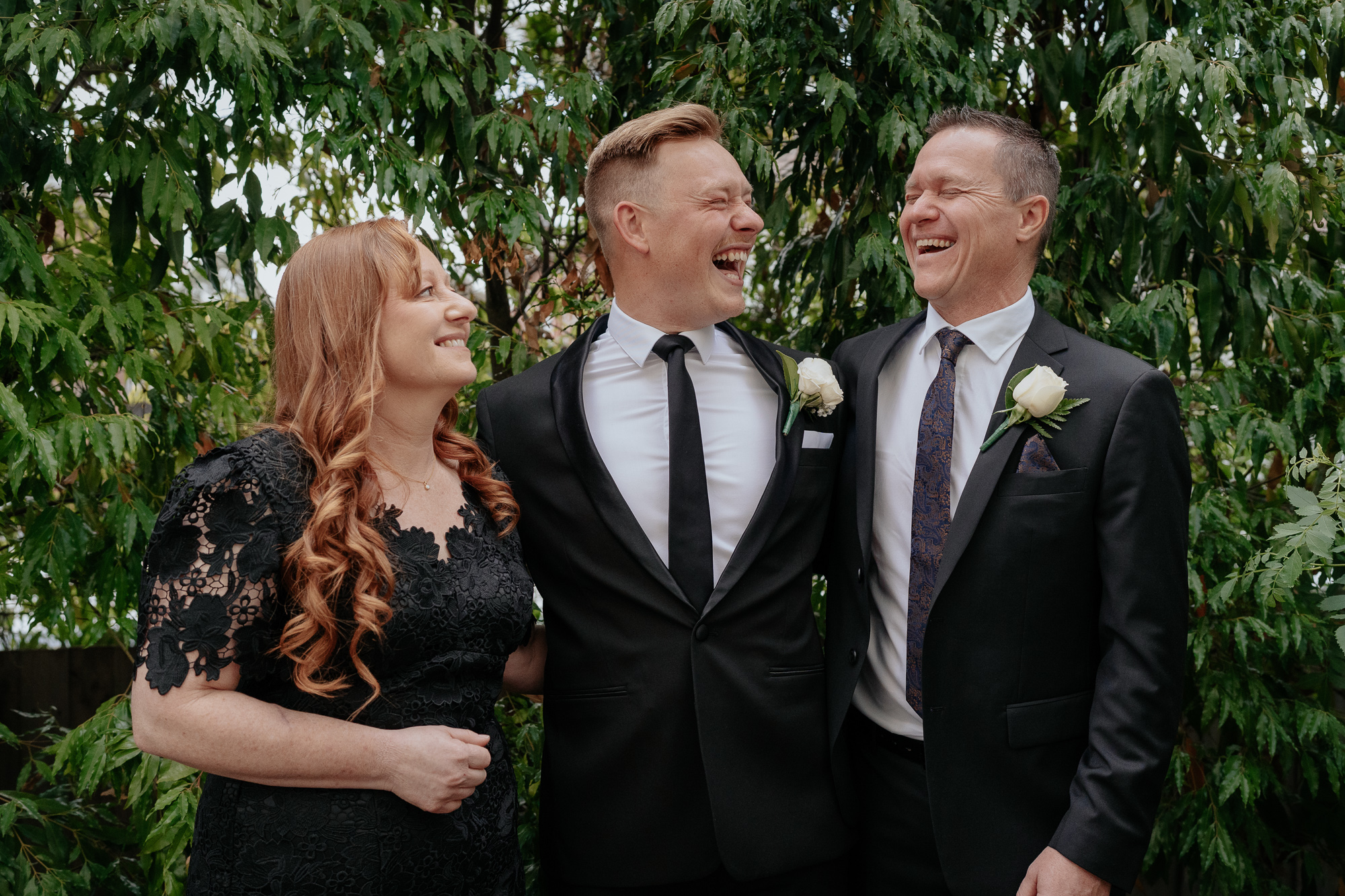 Three people sharing a joyful moment outdoors, two men and a woman, dressed in formal attire, with the two men in tuxedos and the woman in a black lace dress, surrounded by lush green foliage.