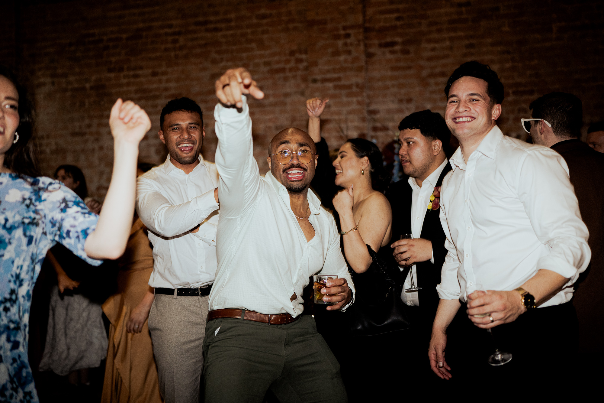 Group of diverse people celebrating at a party or wedding reception, dancing and smiling, with a brick wall in the background. Half Acre South Melbourne Wedding