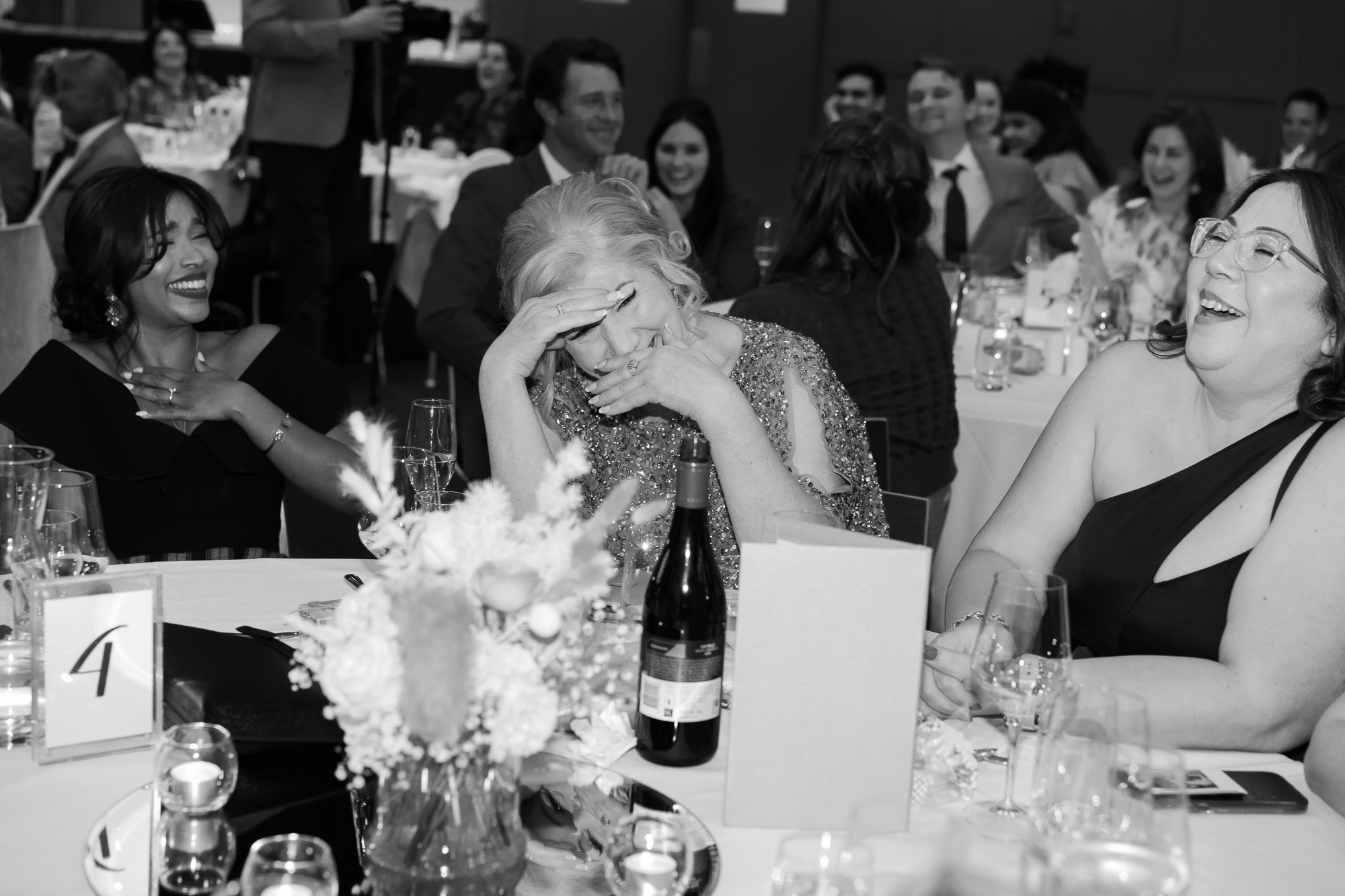 People at a formal event or banquet, laughing and enjoying themselves. There is a woman in the center with her hand on her forehead, seemingly emotional or touched, with others around her smiling or laughing, including women with glasses, a woman wit