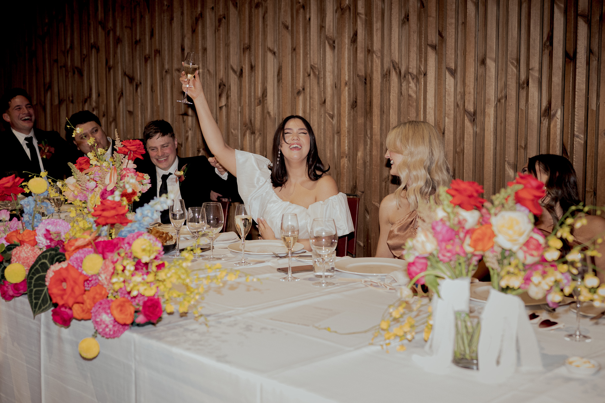 People at a banquet table celebrating, with a woman raising a glass of champagne, surrounded by colorful flowers and wooden wall background. Half Acre Wedding