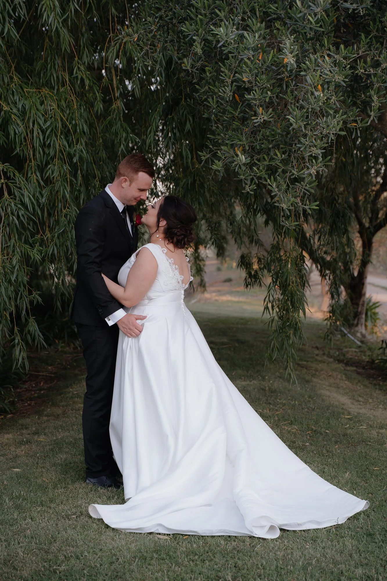 A newlywed couple embraces under a large tree, with the bride in a white wedding gown and the groom in a black suit, in a romantic outdoor setting at Olivehouse at Greendale Grove.
