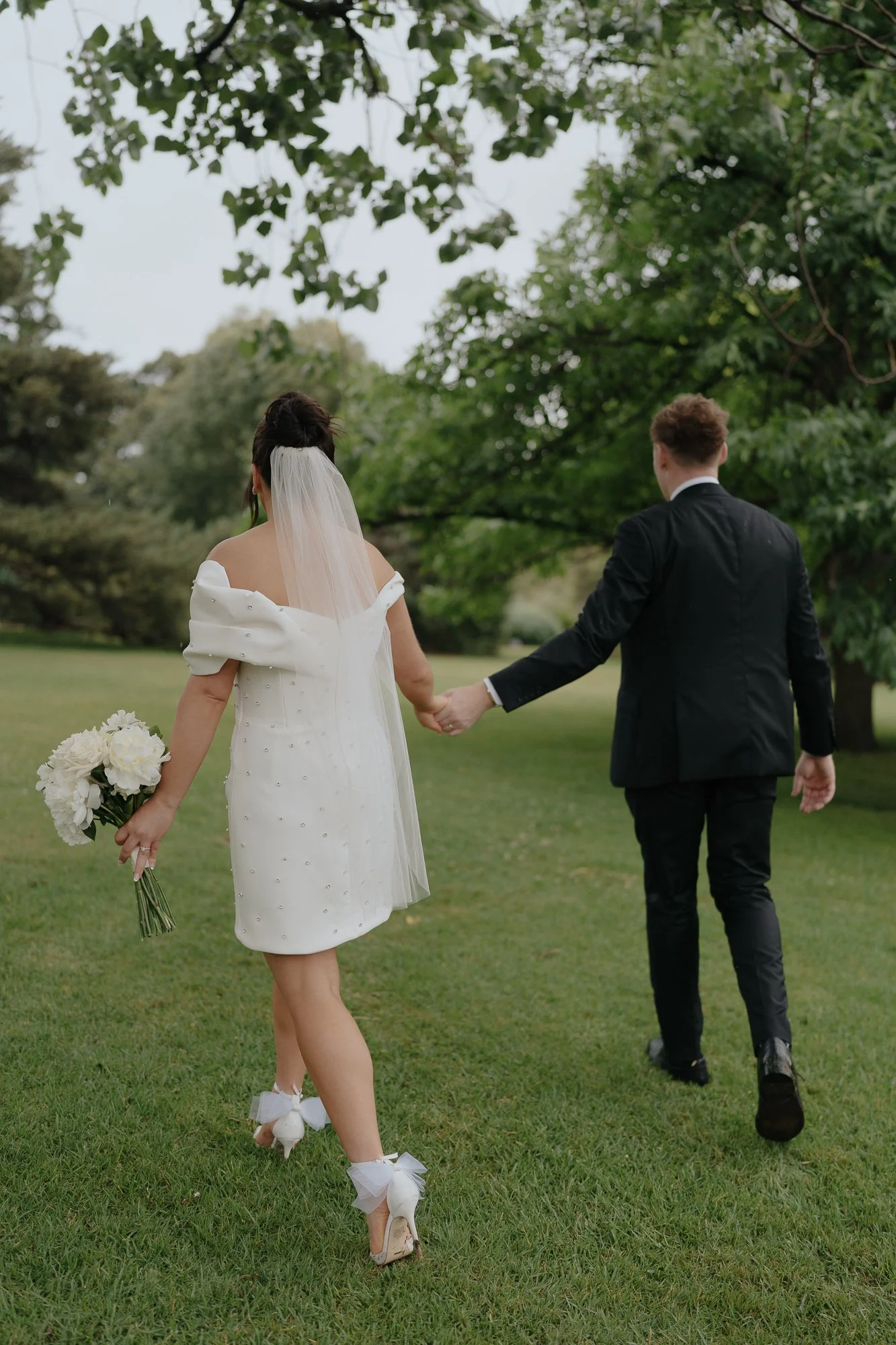 A bride and groom walking hand in hand outdoors on a grassy area with green trees, the bride holding a bouquet of white flowers, wearing a white off-shoulder dress with pom-pom detail and white heels with bows, and the groom wearing a black suit with