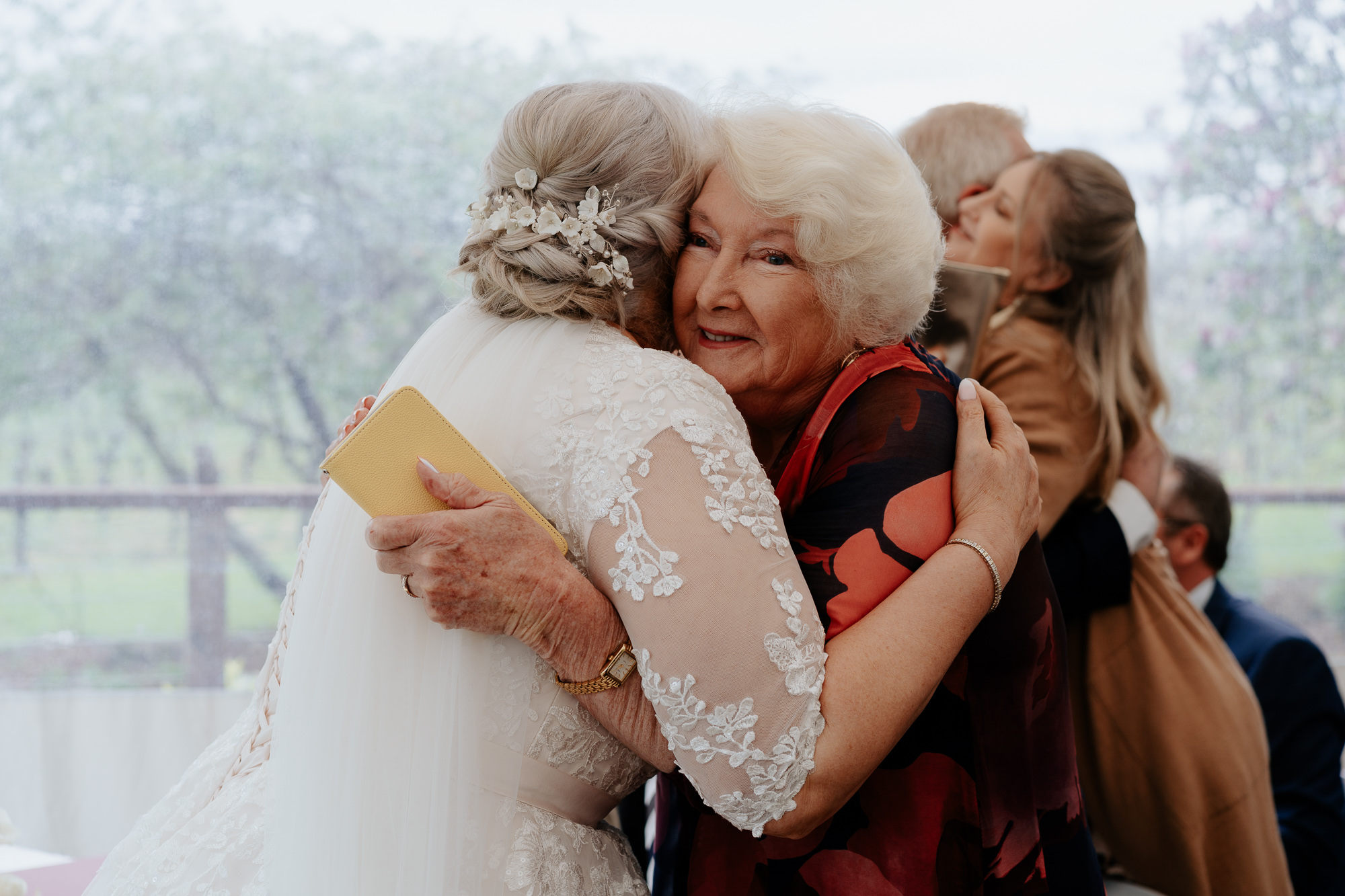 An elderly woman hugging a bride during a wedding celebration, with other guests in the background.
