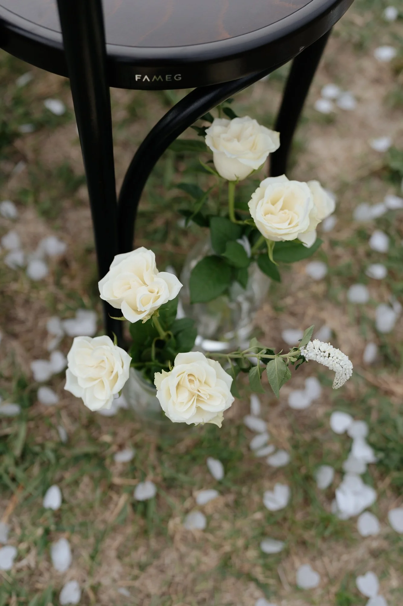 White roses in a glass vase under a brown wooden chair leg, with white flower petals scattered on the ground at the Vines on Avon.