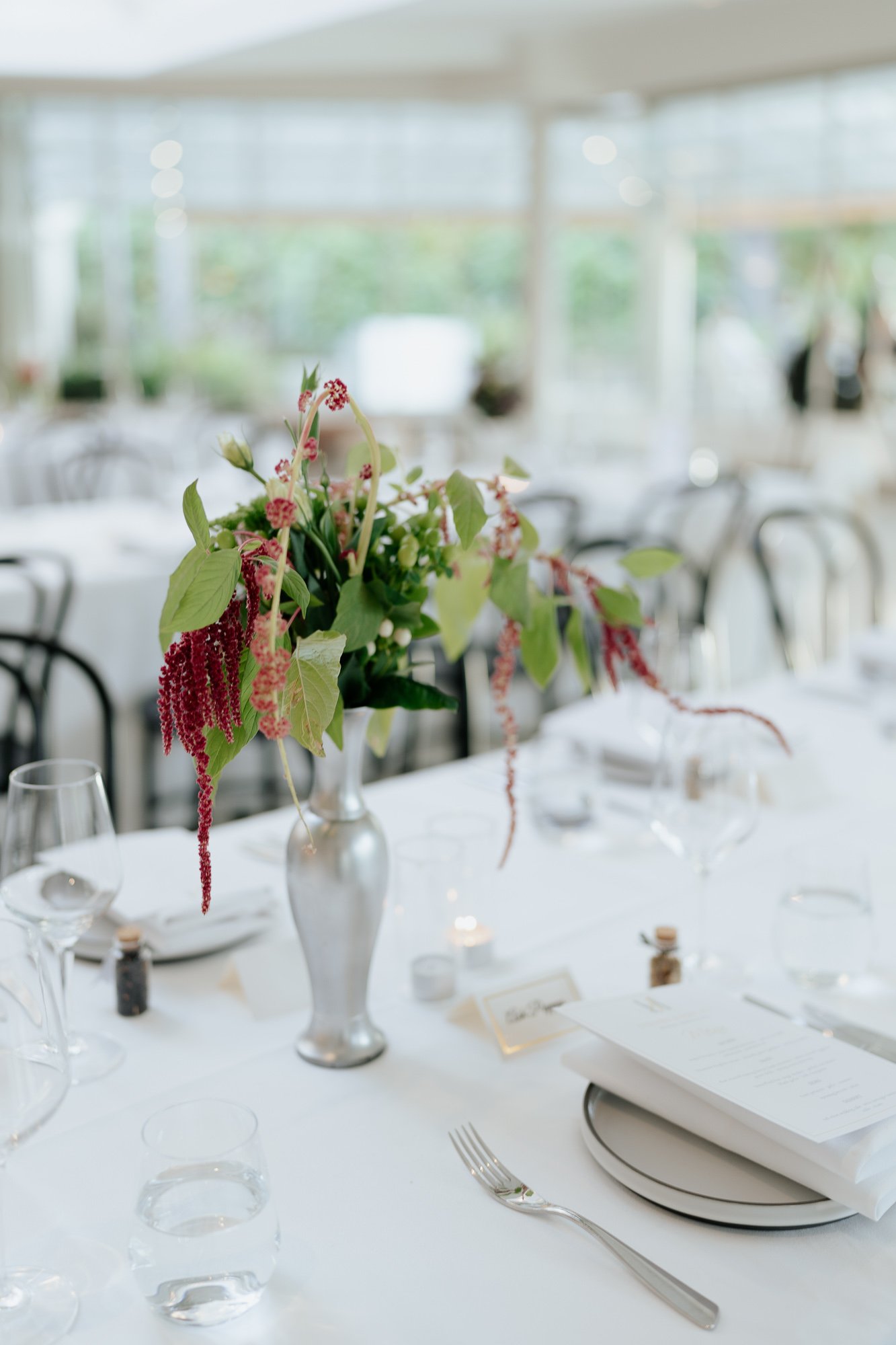 Elegant table setting with a silver vase holding pink and green floral arrangement, glassware, menu, and place card on a white tablecloth in a well-lit, modern venue.