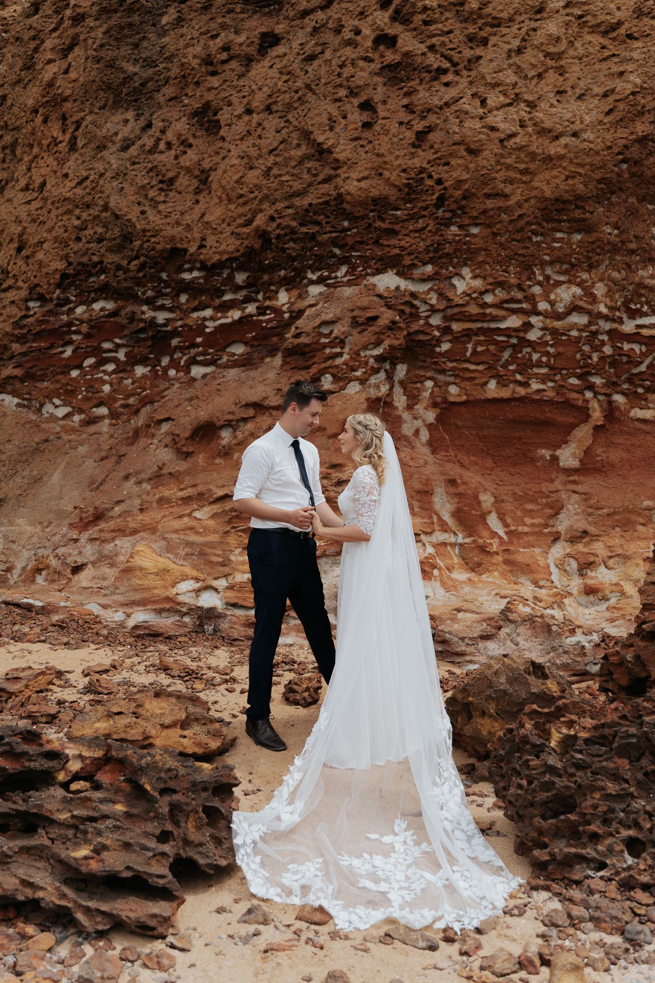 A bride and groom standing on a sandy area with large rocky formations behind them, holding hands and gazing at each other. The bride is wearing a white lace wedding dress with a long train, and the groom is dressed in a white shirt and dark pants.