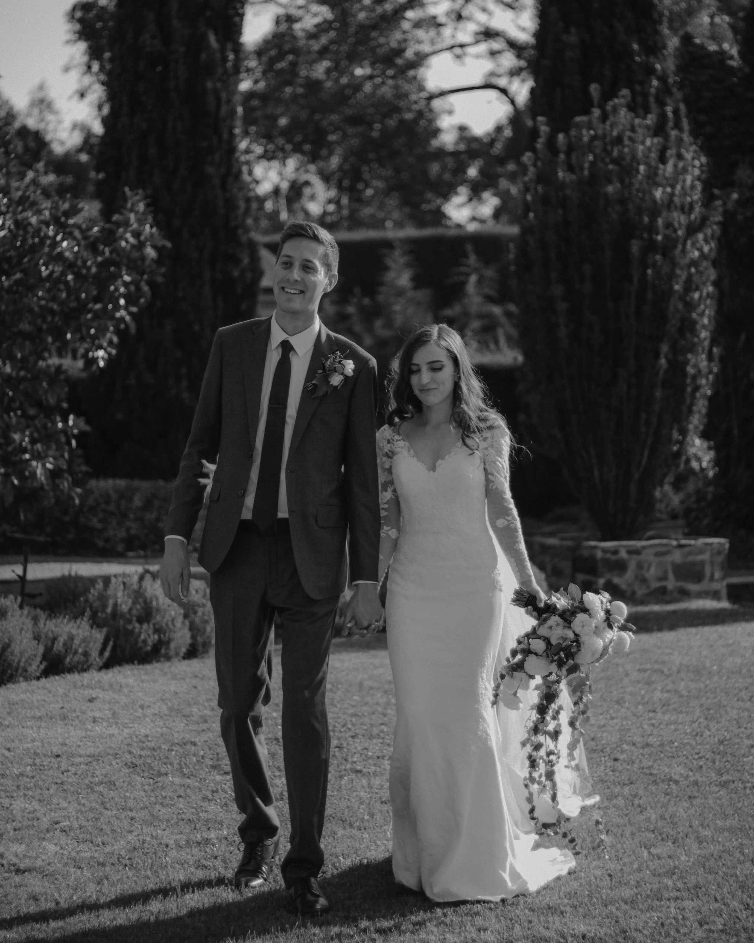 A black and white photo of a bride and groom walking outdoors on their wedding day, holding hands, with trees and shrubs in the background.