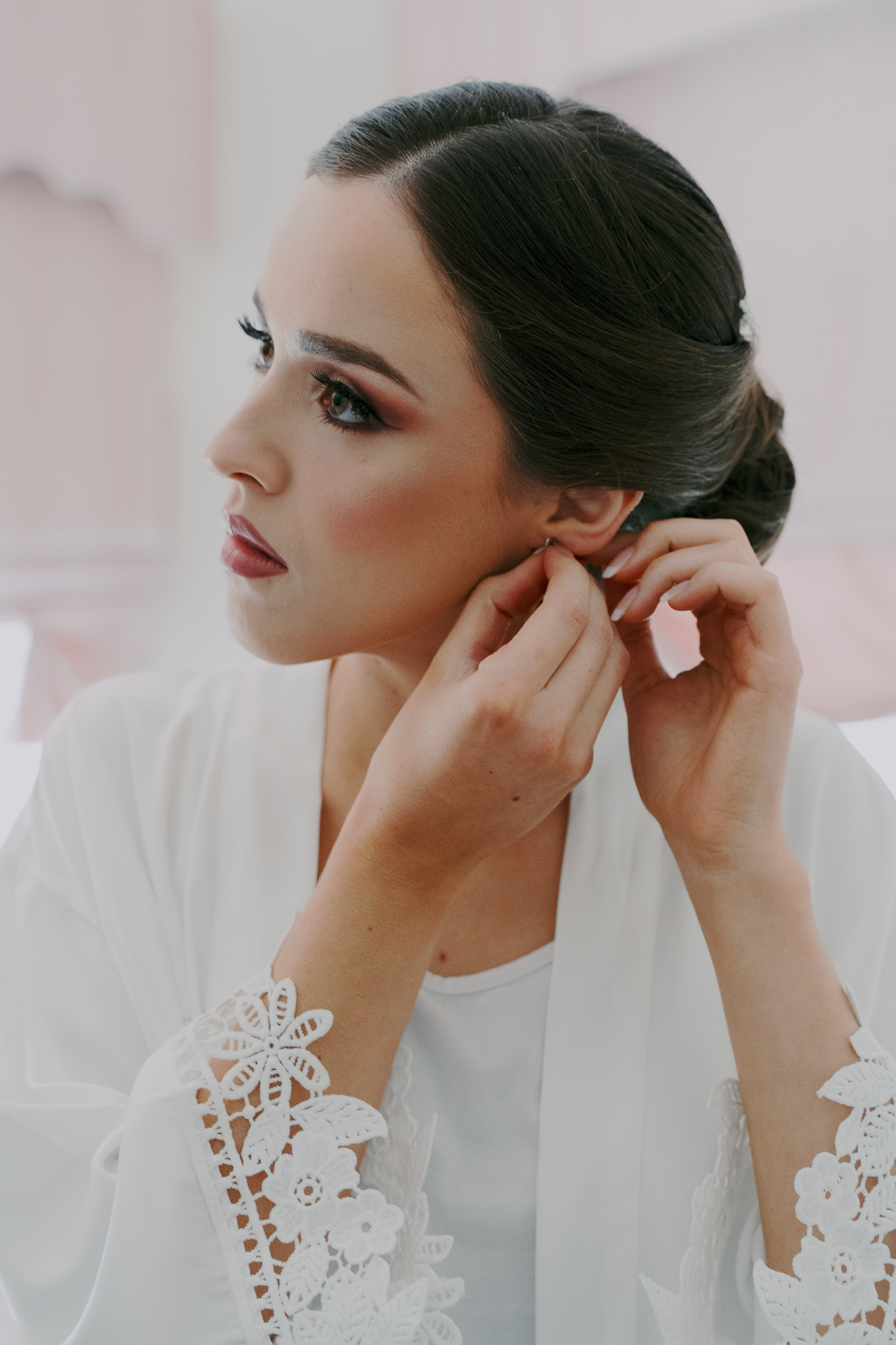 A woman with dark hair styled in an elegant updo puts on a pair of earrings. She wears a white top with lace detailing on the sleeves.