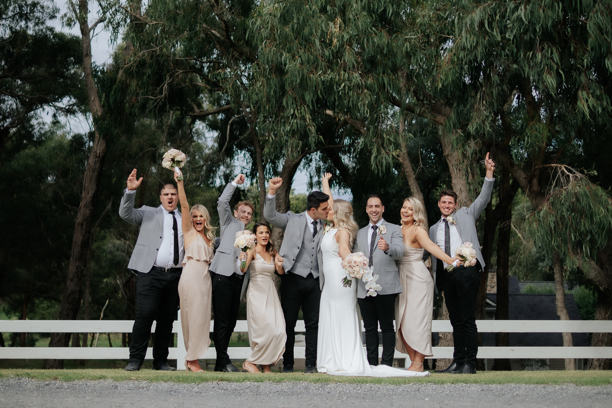 Smiling bride and groom kissing surrounded by wedding party celebrating outdoors.