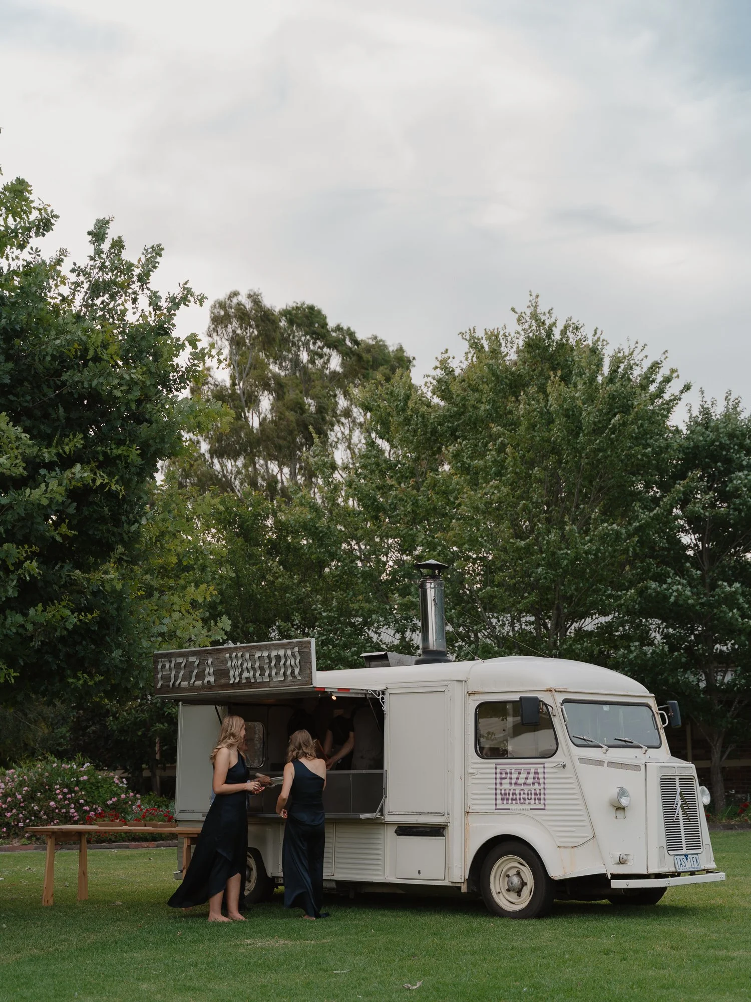 A white pizza food truck with a sign that says "Pizza Wagon" on the side, parked on a grassy area surrounded by trees. Two women in formal dresses are standing in front of the truck, ordering food.
