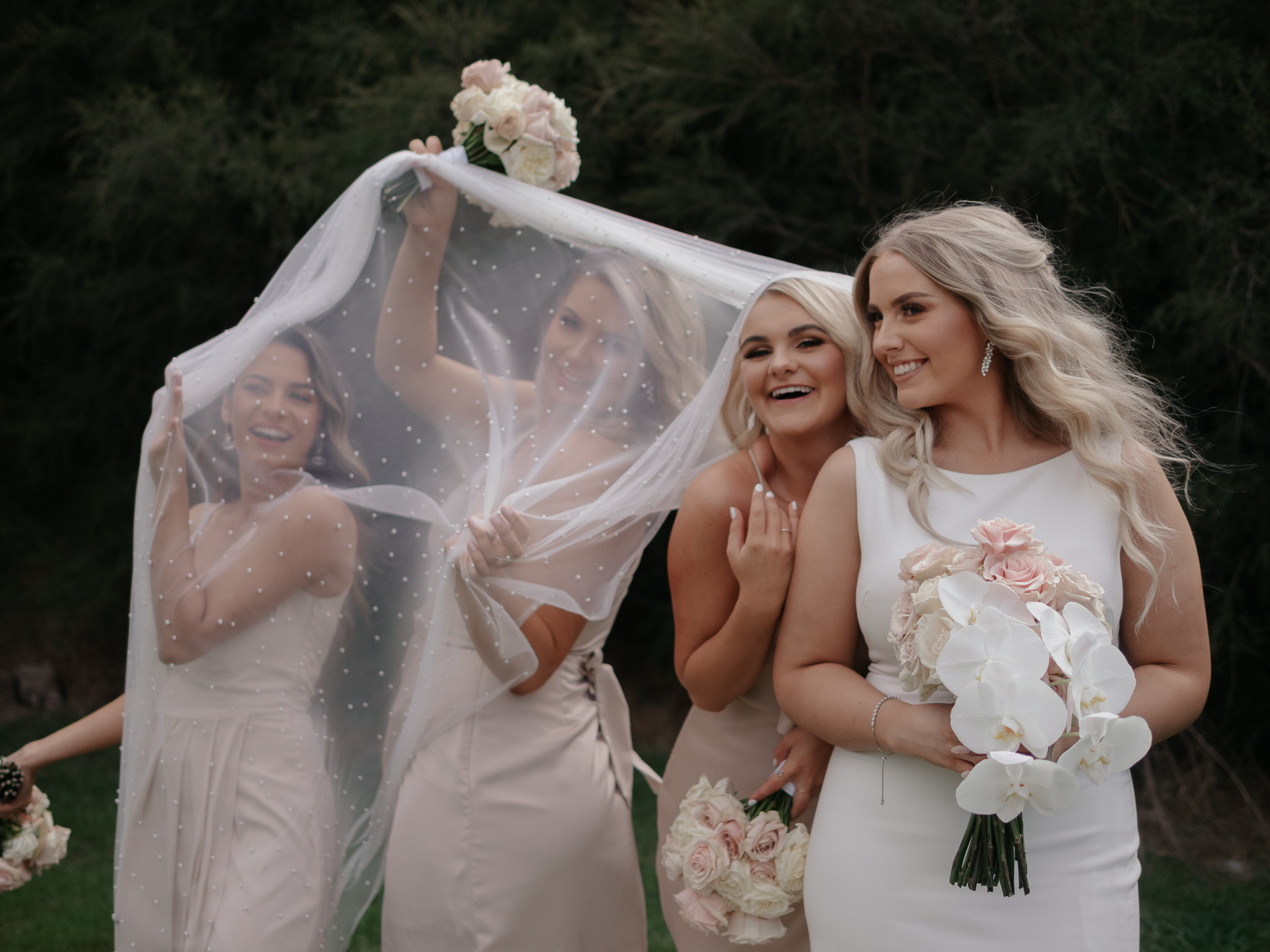 Four women in wedding dresses pose outdoors; one bride holds a bouquet of white flowers with pink roses, with her veil and friends behind her smiling. Bramleigh Estate Wedding