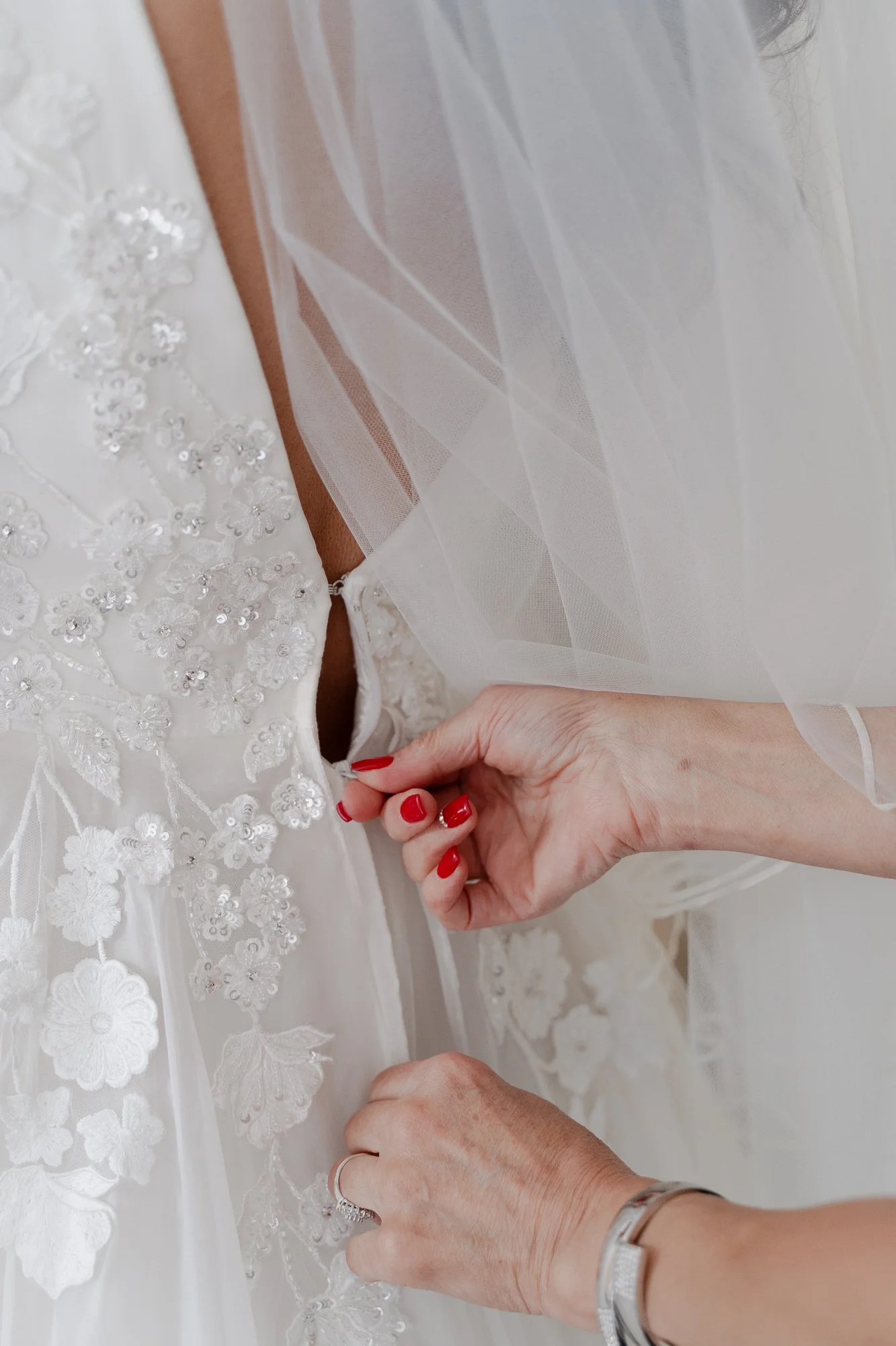 Close-up of a bride's hands buttoning the back of her wedding dress, which features lace embroidery and floral appliqué details, while wearing a silver bracelet and red nail polish.
