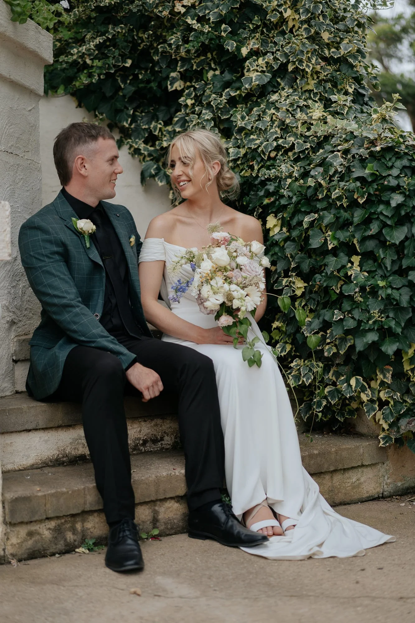 Gippsland Wedding Photography. A bride and groom sitting together outdoors at Amarti at the Heritage Listed Cowwarr Butter Factory, smiling at each other, with the bride holding a bouquet of flowers, against a background of green ivy.