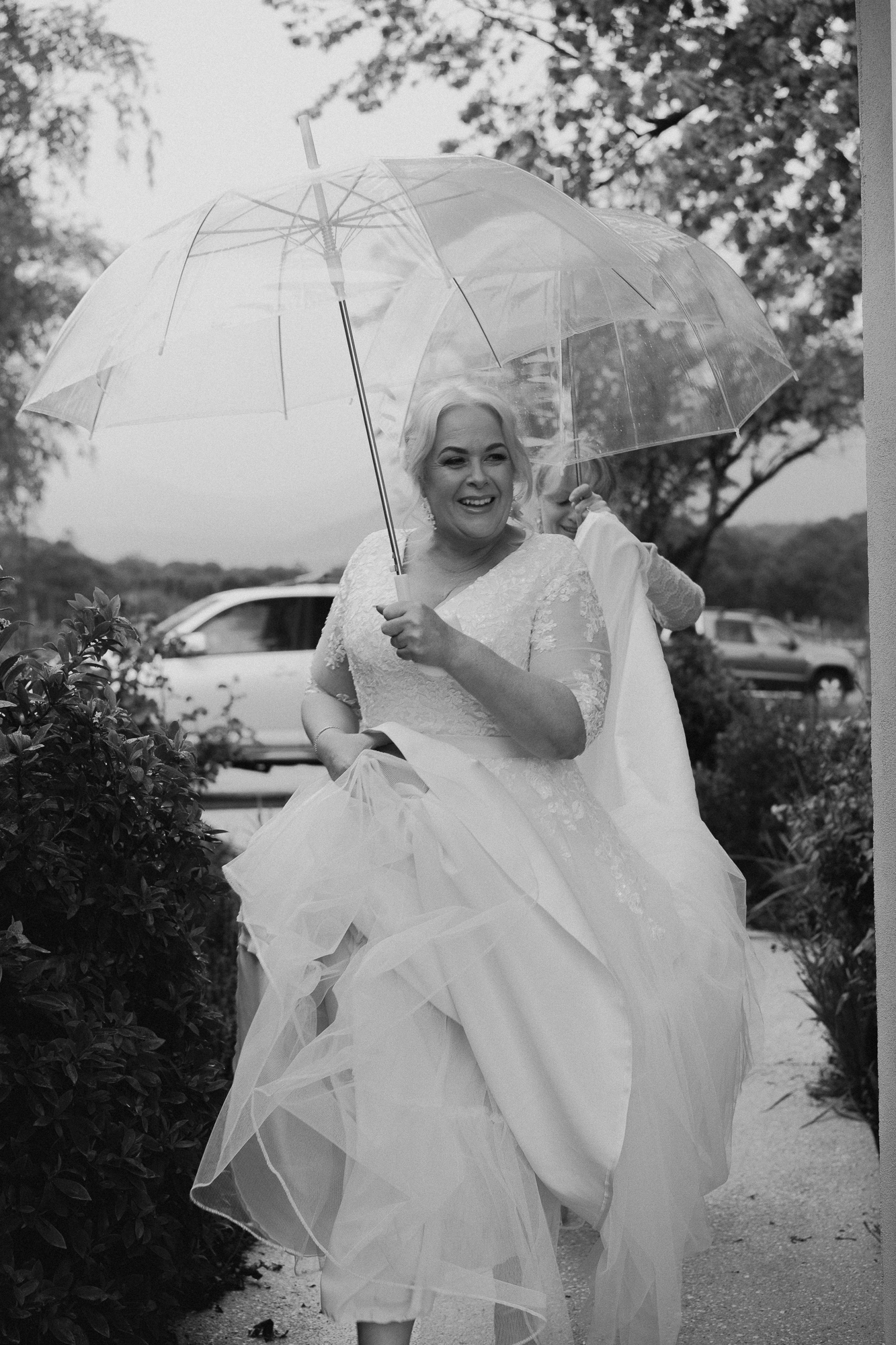 A bride in a wedding dress holding a transparent umbrella, smiling, with a person behind her holding her dress, outdoors with trees and parked cars.
