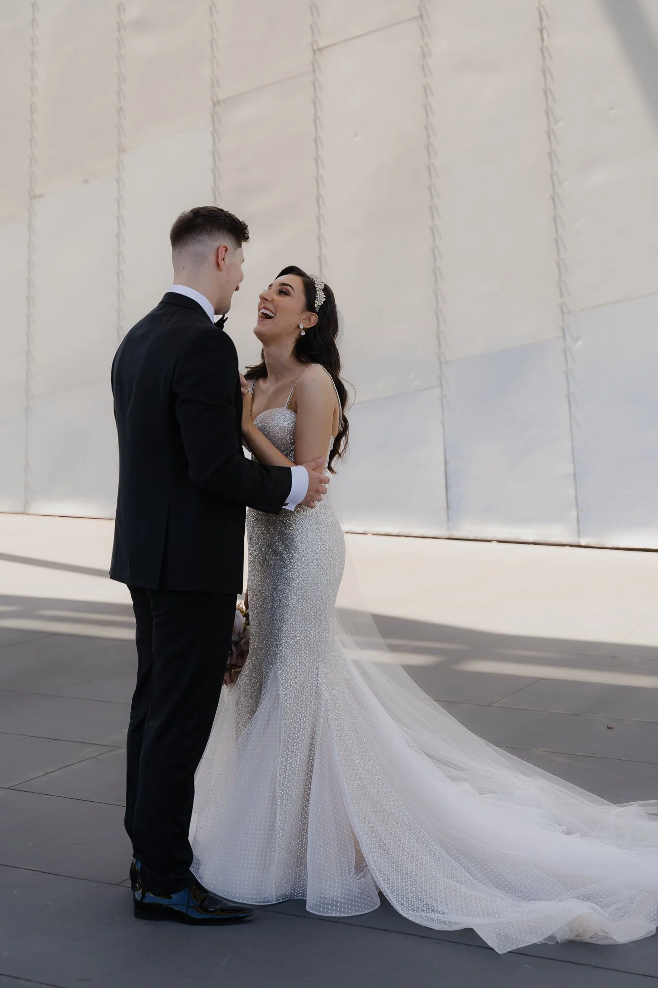 A bride and groom sharing a joyful moment outdoors, with the bride in a sparkling wedding gown and the groom in a black tuxedo.