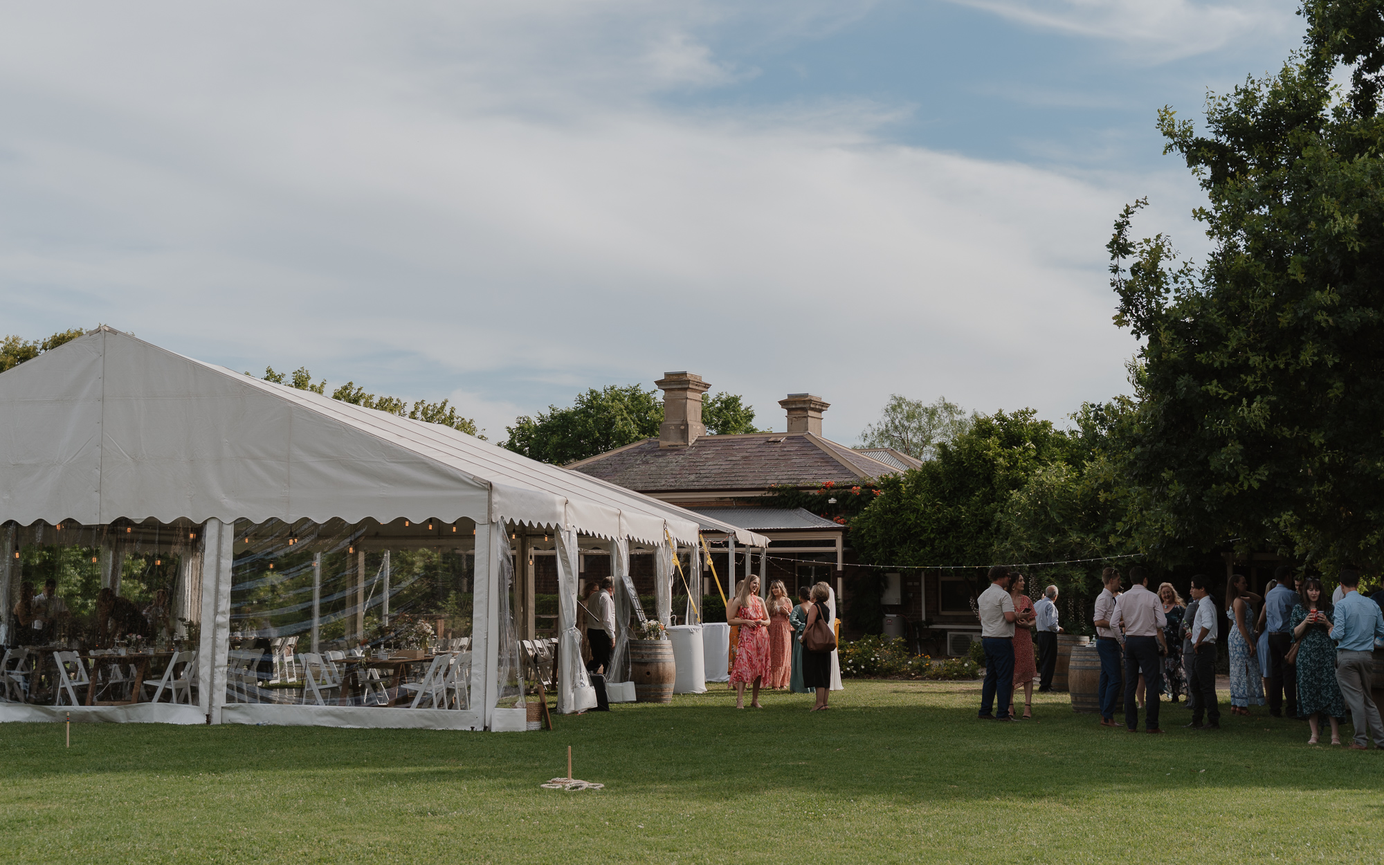 People socializing outdoors at a wedding reception or event, with a large white tent, a building with chimneys, and trees in a grassy backyard.