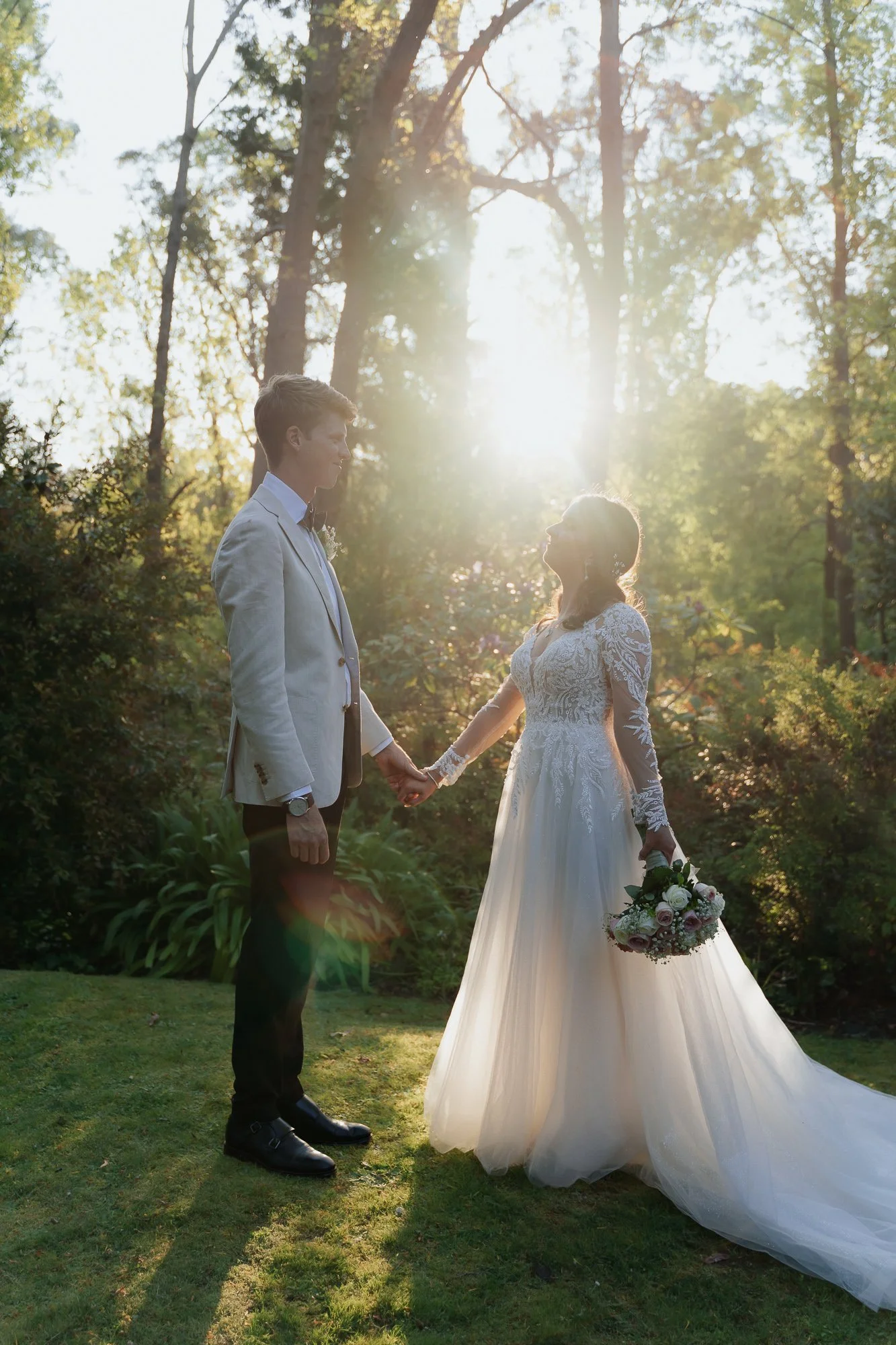 A bride and groom holding hands outdoors during sunset at Chateau Wyuna, with trees in the background. The woman is wearing a long white wedding dress and holding a bouquet, while the man is dressed in a light-colored suit.