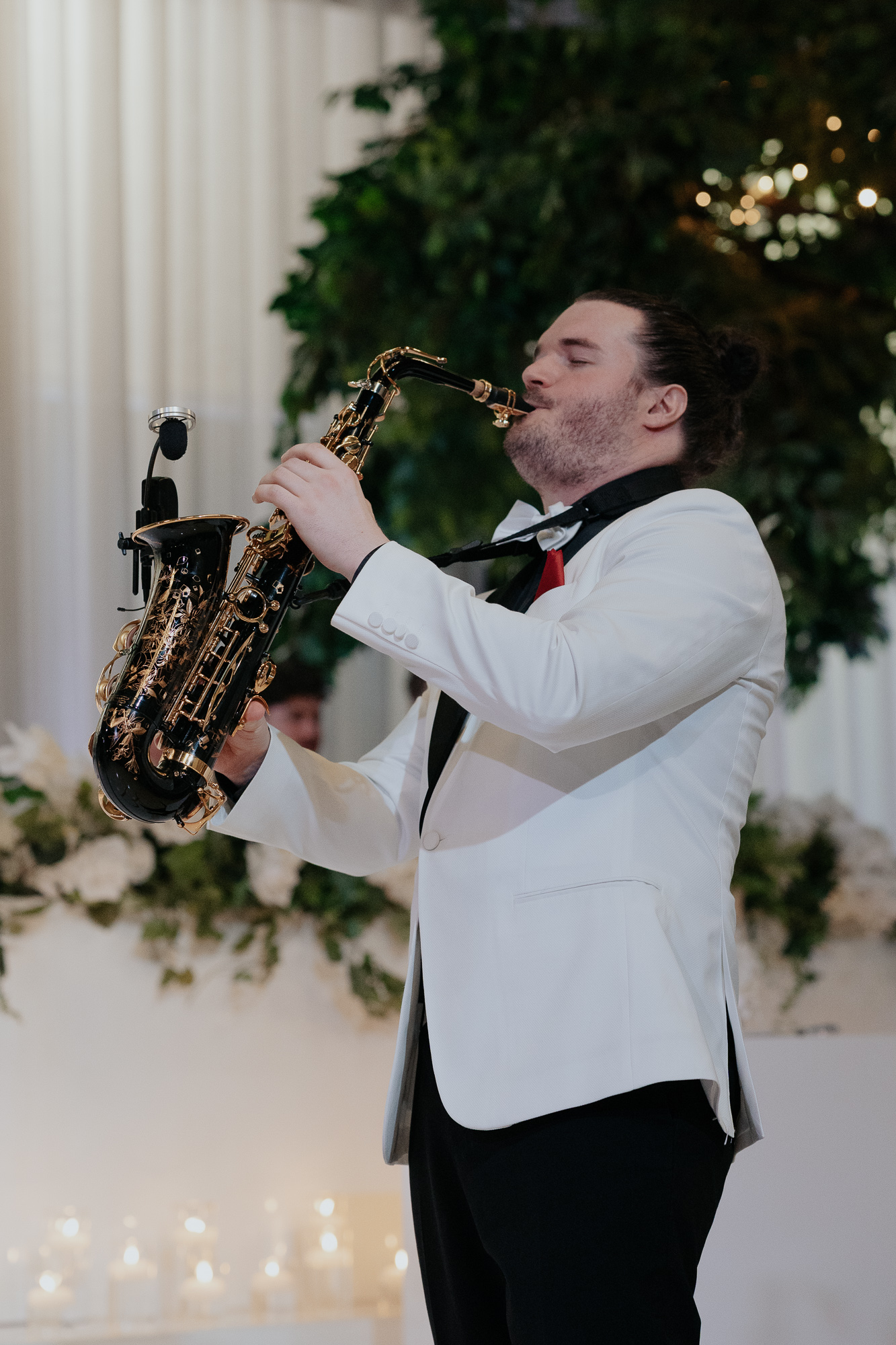 A man in a white tuxedo jacket and black pants playing a saxophone, at an event with elegant floral decorations and candles in the background.