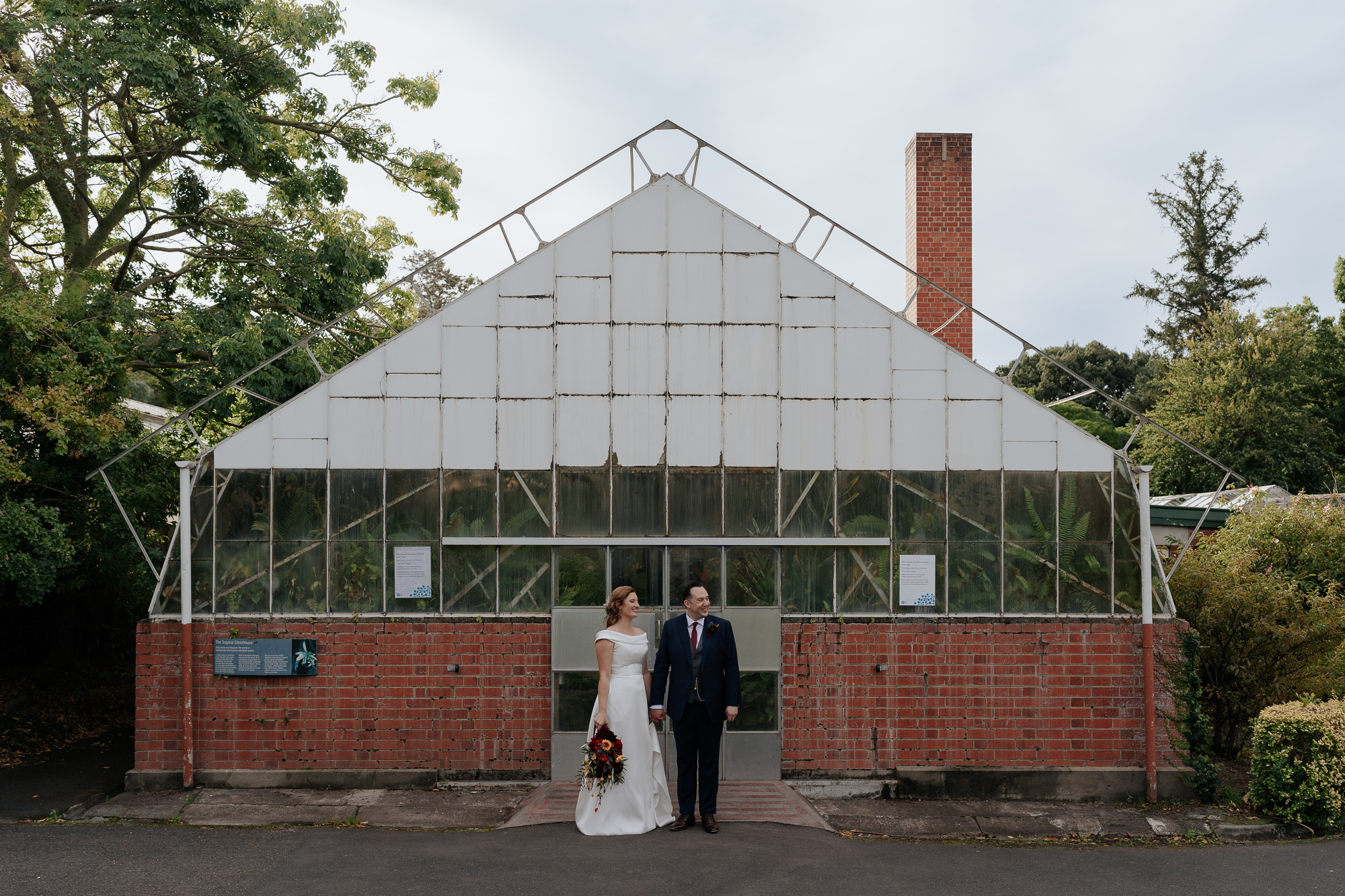 A newlywed couple holding hands standing outside a vintage greenhouse with brick foundation and glass panels, trees in the background.
