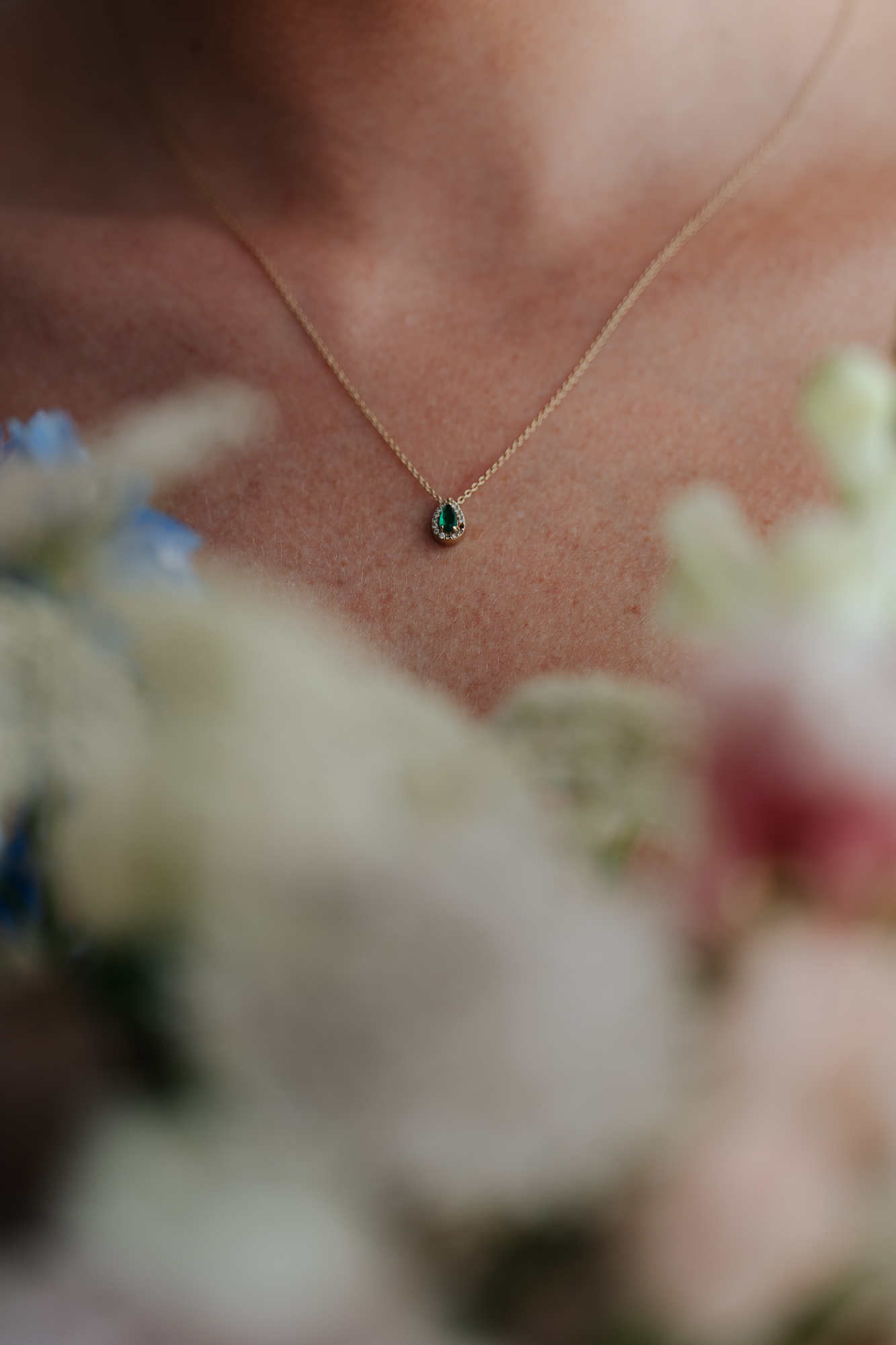 Close-up of a person wearing a gold necklace with a small teardrop-shaped dark green gemstone pendant, surrounded by blurred flowers in the foreground. Amarti Wedding Venue.