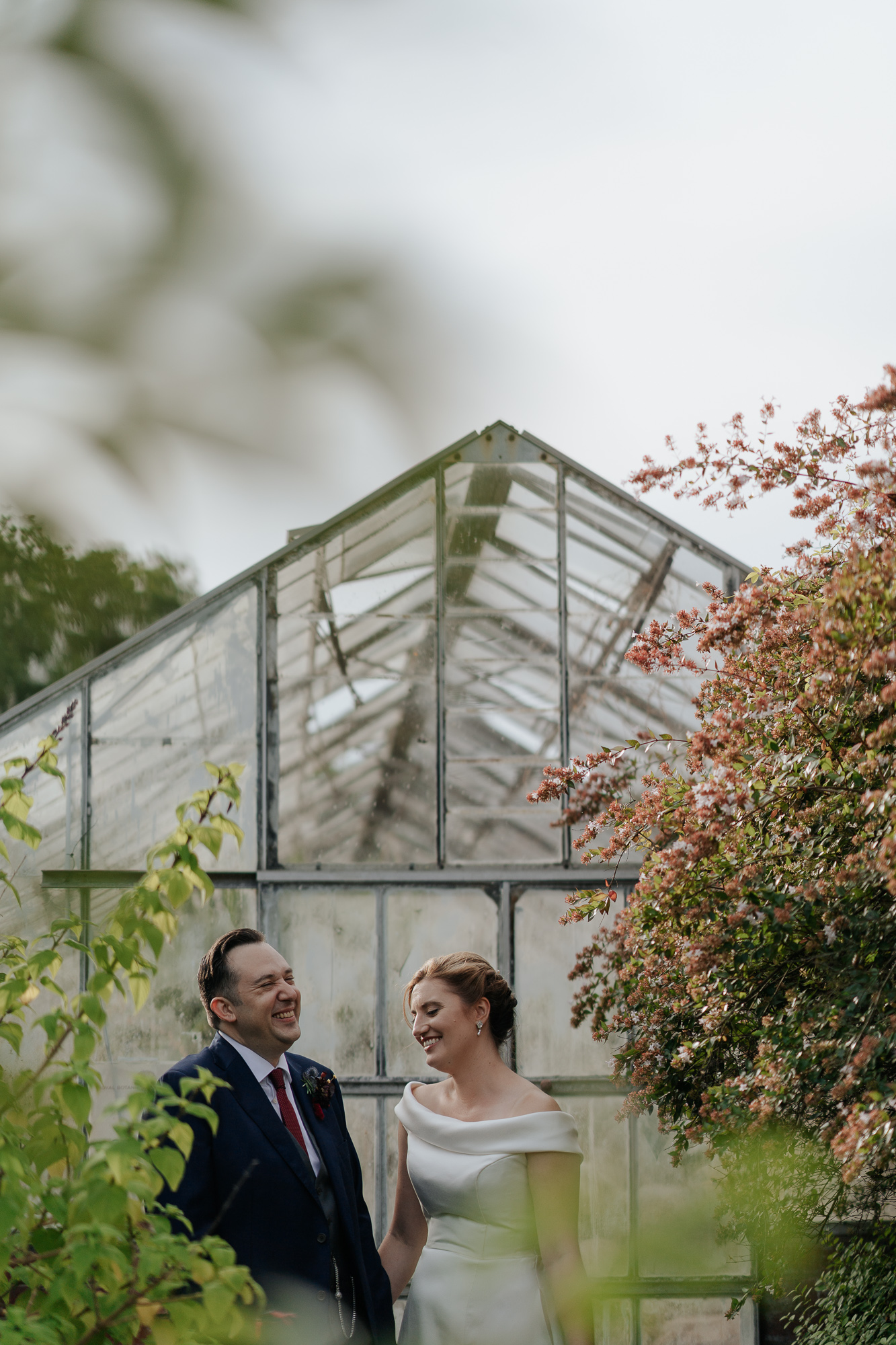 A bride and groom standing together smiling in a greenhouse, surrounded by green plants and pink flowering trees, during their wedding day. Royal Botanical Gardens Melbourne