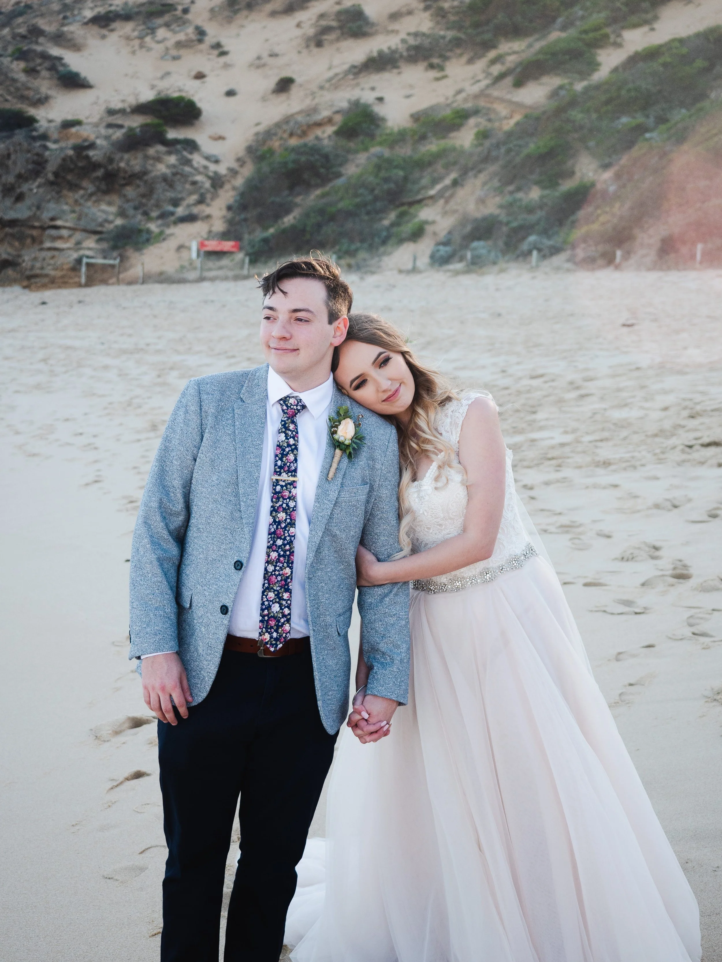 Mornington Peninsula Wedding Photography. A young couple in wedding attire holding hands on a sandy beach on the Mornington Peninsula, with the woman resting her head on the man's shoulder as they pose for a photo.