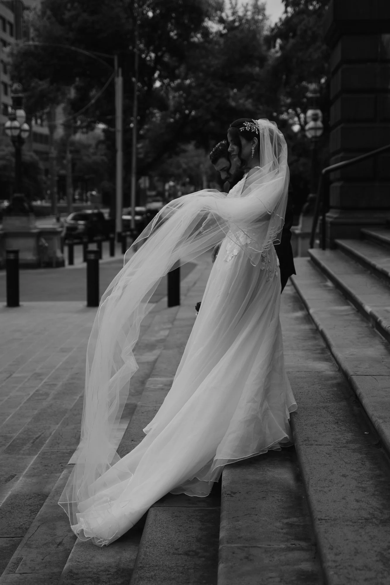Black and white photo of a bride and groom on the steps of a building, with the bride wearing a long veil and dress, and the groom in a suit. The bride is holding up her veil as they share a moment in an urban setting.