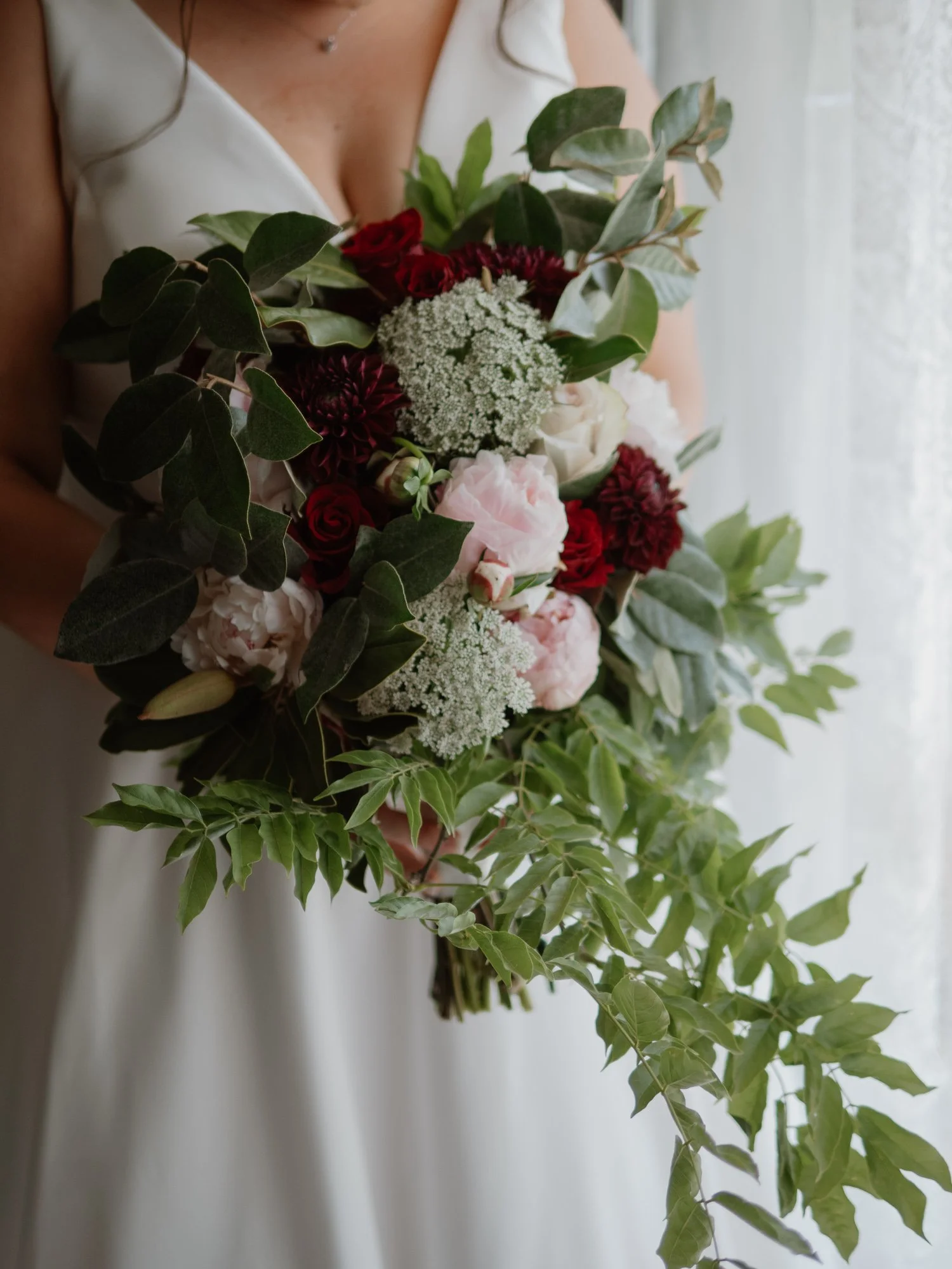 A woman in a white dress holding a large bouquet of red, pink, and white flowers with greenery, near a window with curtains.