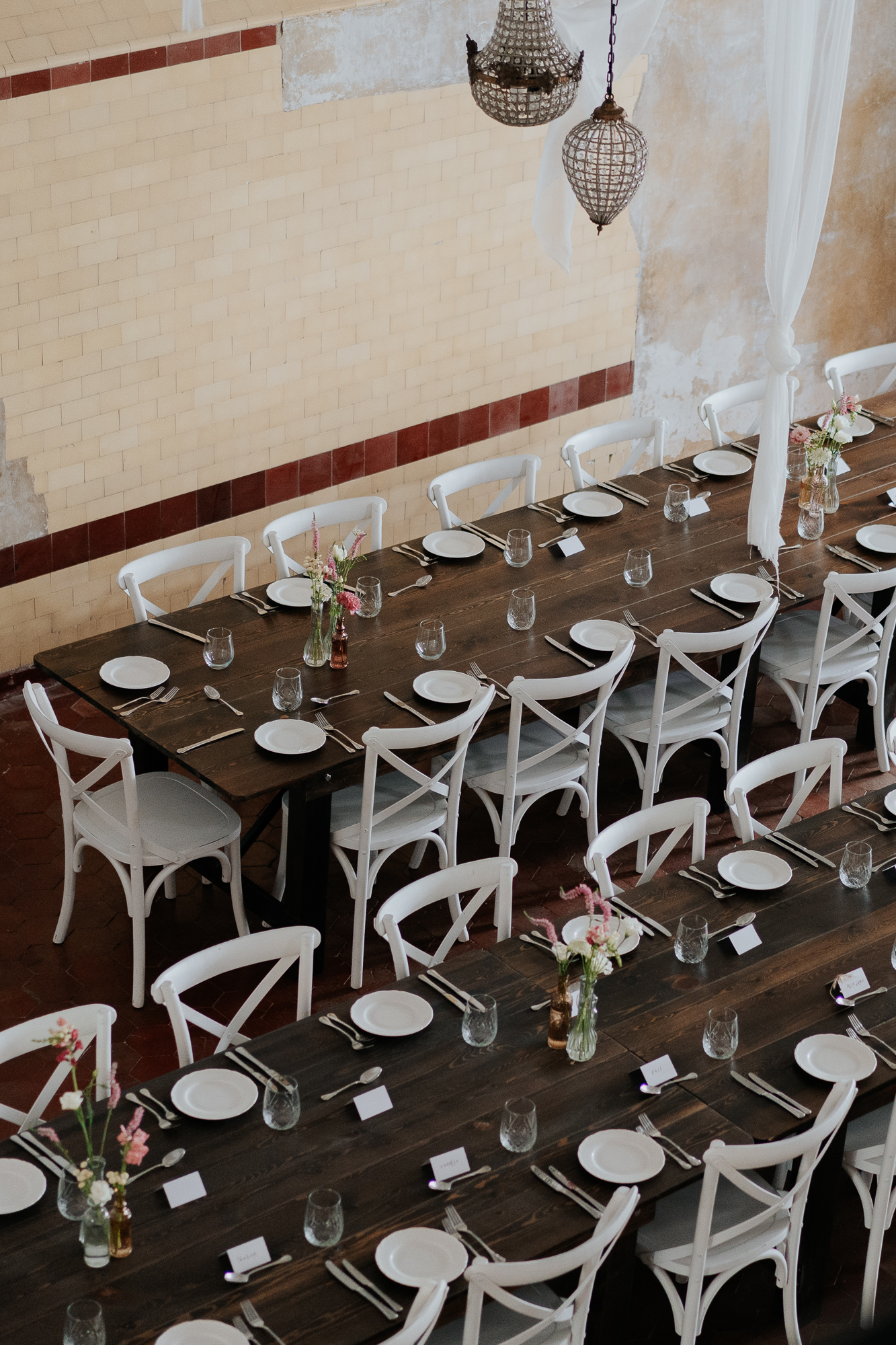 Long wooden dining table set for a formal event, with white chairs, plates, glasses, and small flower arrangements, in a rustic-style room with wall sconces and hanging lights.
