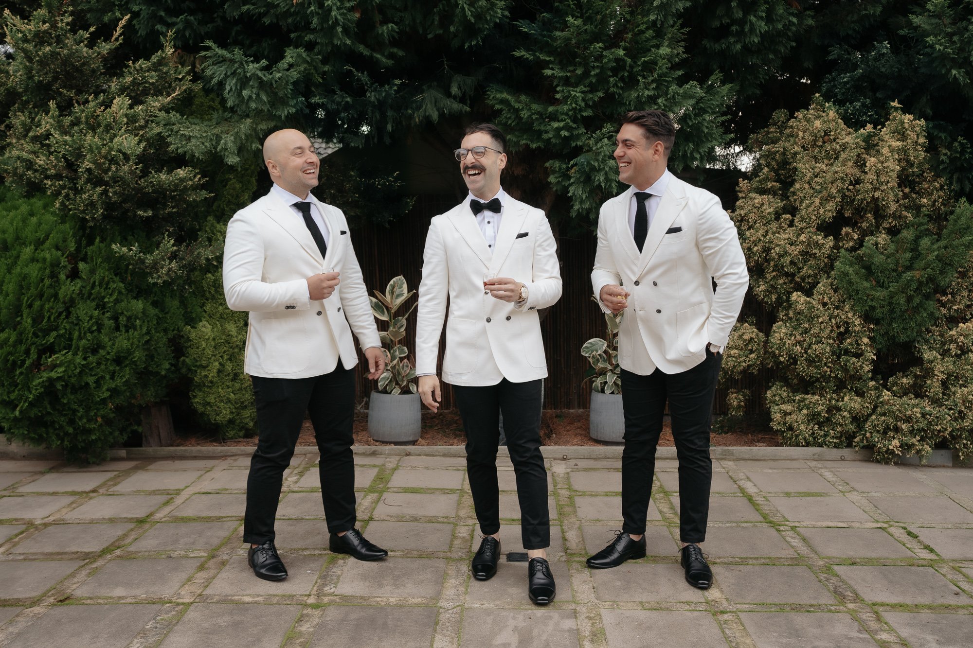 Three men dressed in white tuxedos and black pants, standing on a paved patio with green trees and potted plants behind them, laughing and conversing, each holding a glass.