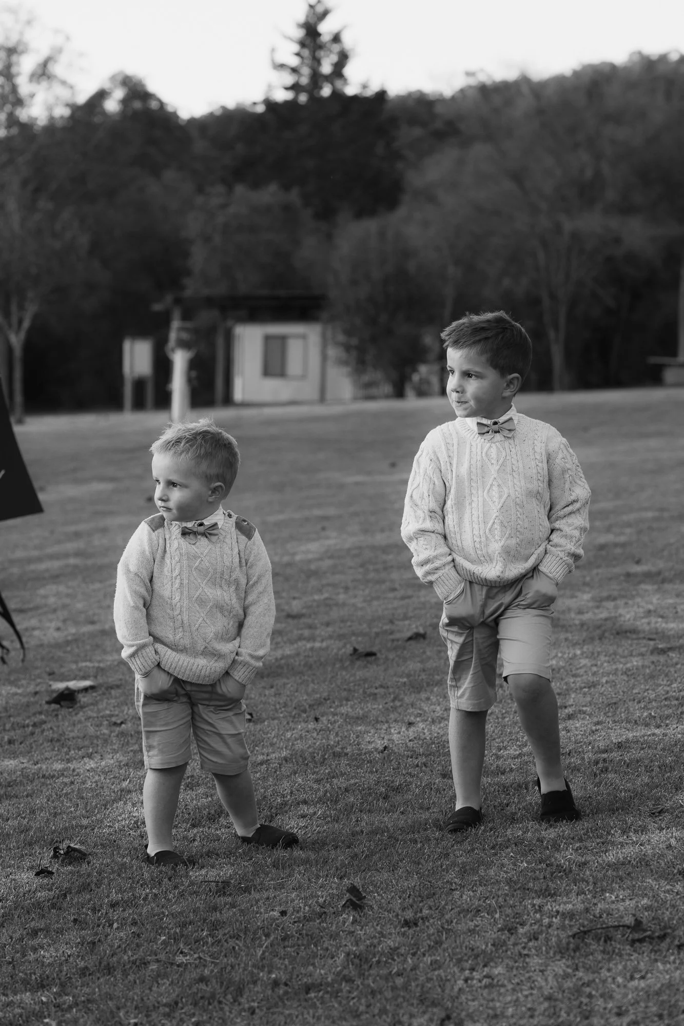 Two young boys dressed in sweaters, shorts, and bow ties, standing on grass in an outdoor setting, with trees and a building in the background.