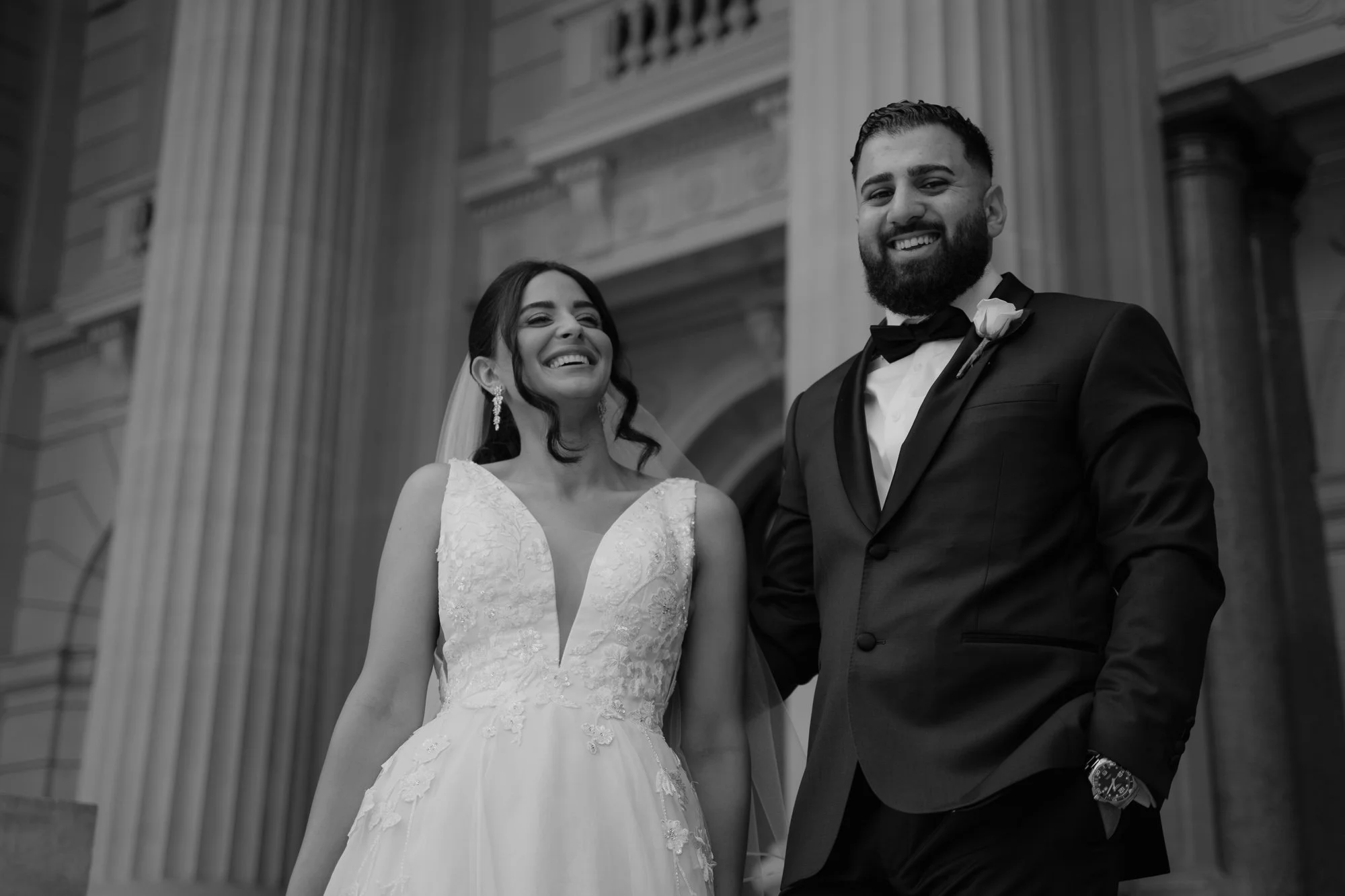 A bride and groom smiling and holding hands outside a historic building during their wedding.