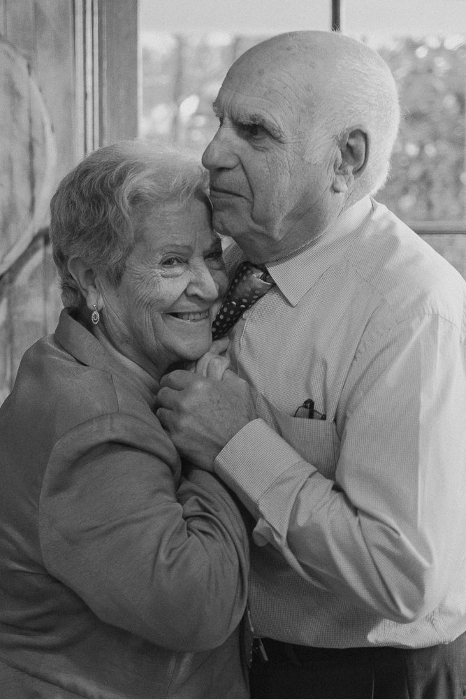 An elderly couple dancing indoors, the woman smiling and holding onto the man's hand, both dressed in formal attire.