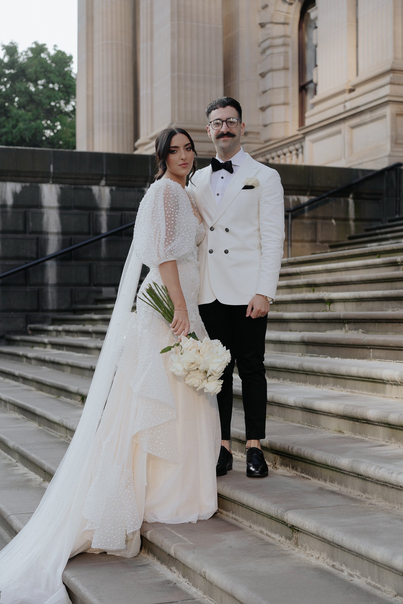 Bride and groom standing on stone steps outside a historic building, dressed in wedding attire; she is holding a bouquet of white flowers, and he is dressed in a white tuxedo jacket with black pants.