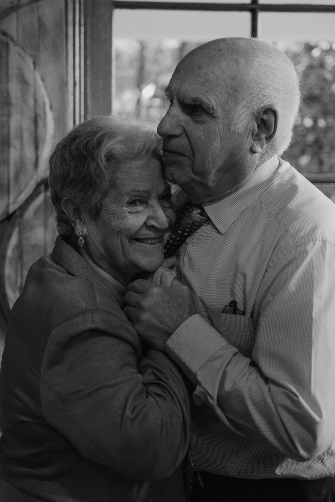 An elderly man and woman sharing a joyful embrace, smiling warmly. The man is tenderly holding the woman and they are close in a cozy indoor setting.