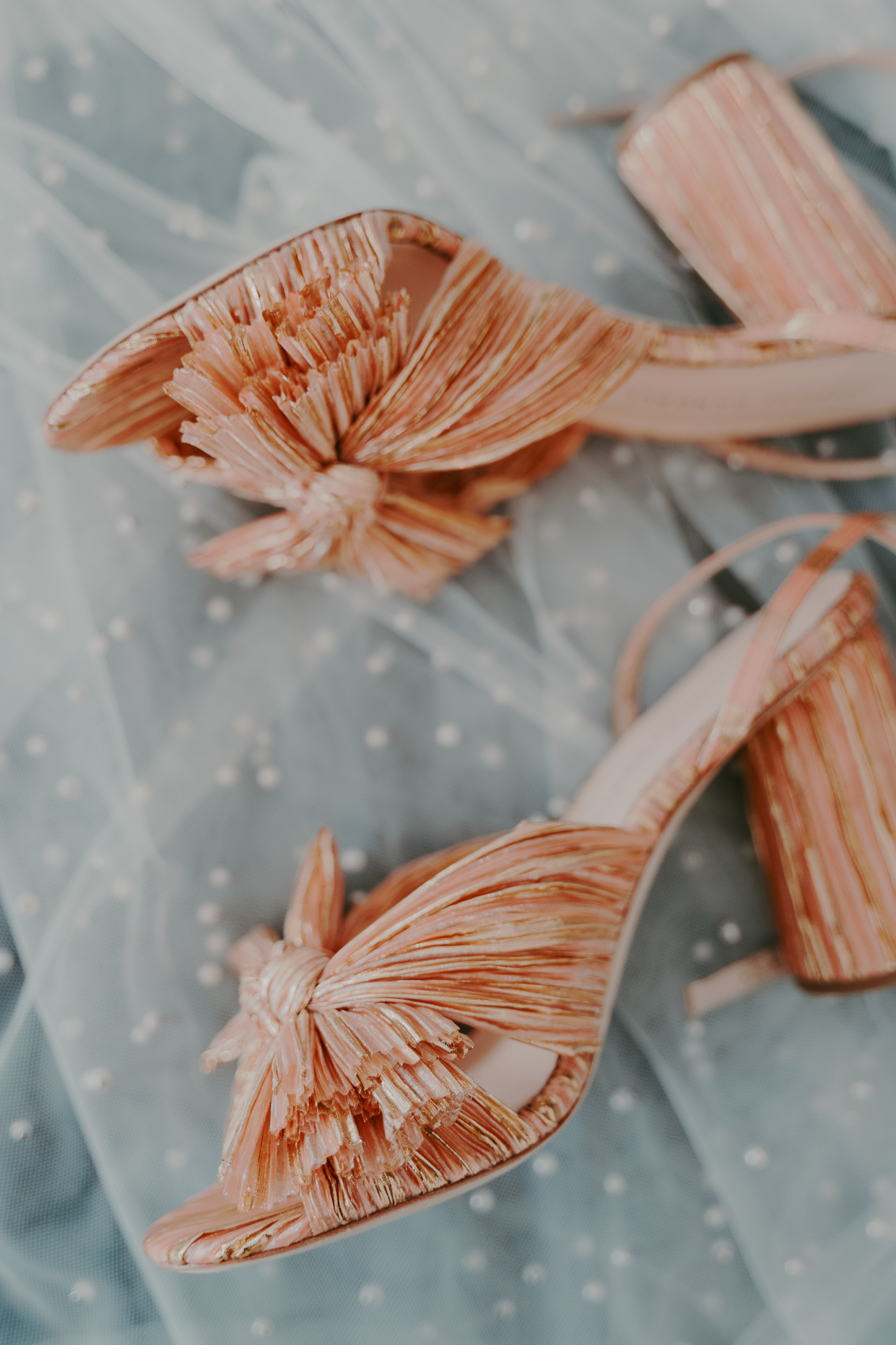 Close-up of pink and orange striped high-heeled Loeffler Randall shoes with fringed bows, resting on sheer blue fabric with white polka dots.