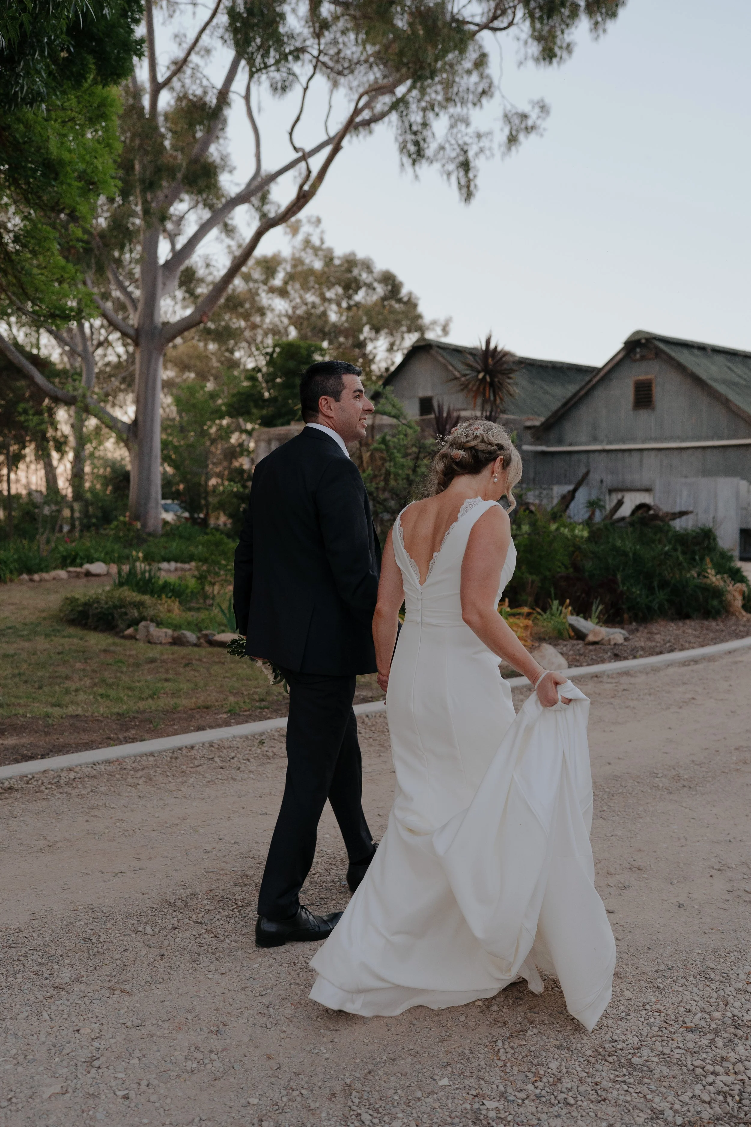 A bride in a white wedding dress and a groom in a black suit walking hand in hand outdoors on a dirt path, with trees and old wooden buildings in the background. On the grounds of Buller Wines, Rutherglen.